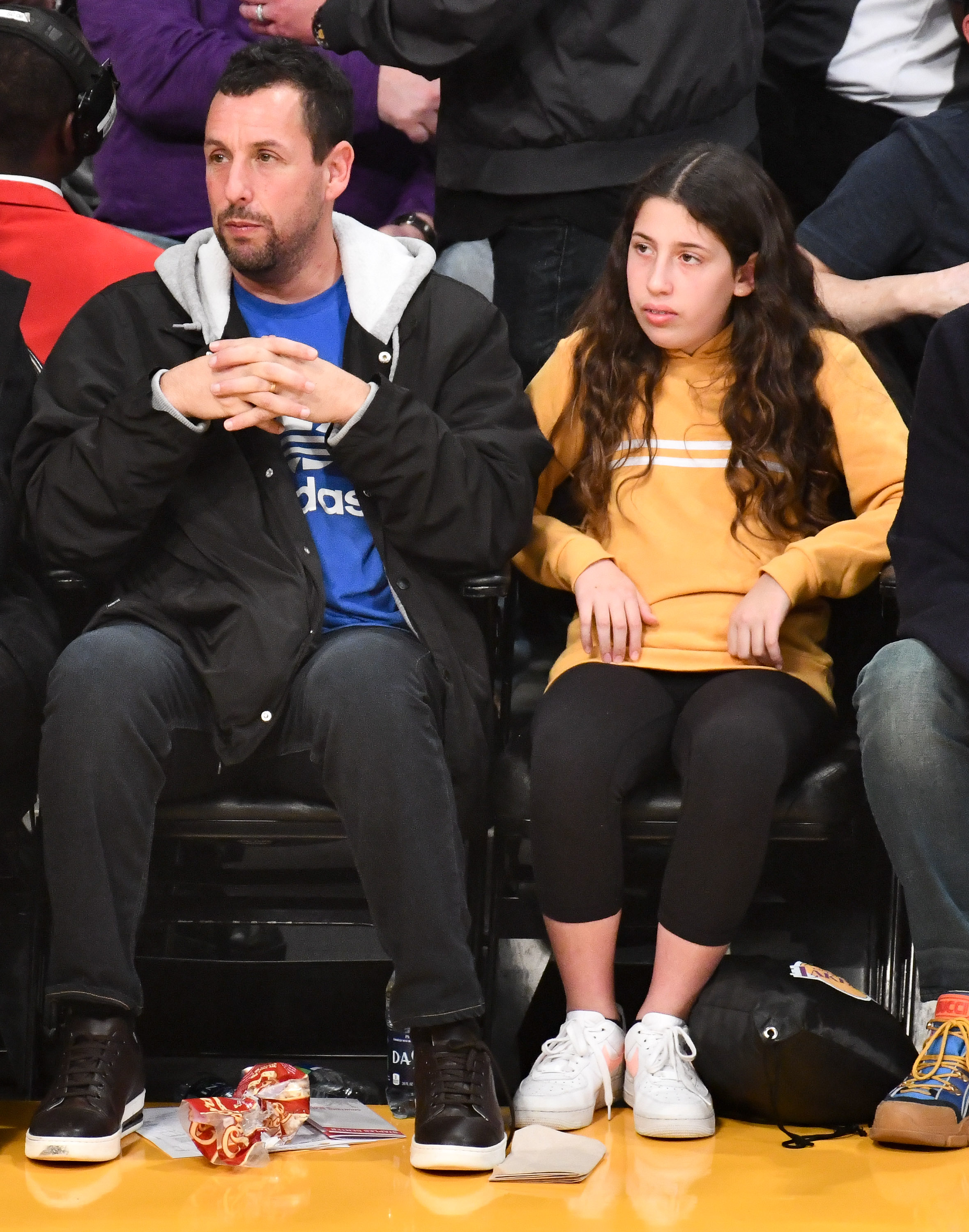 Adam Sandler sits courtside beside Sadie Madison Sandler, both locked into the game as the action unfolds just feet away. In a casual hoodie-and-sneakers look, he leans forward with hands clasped, while Sadie mirrors the focus in a mustard sweatshirt and white kicks.