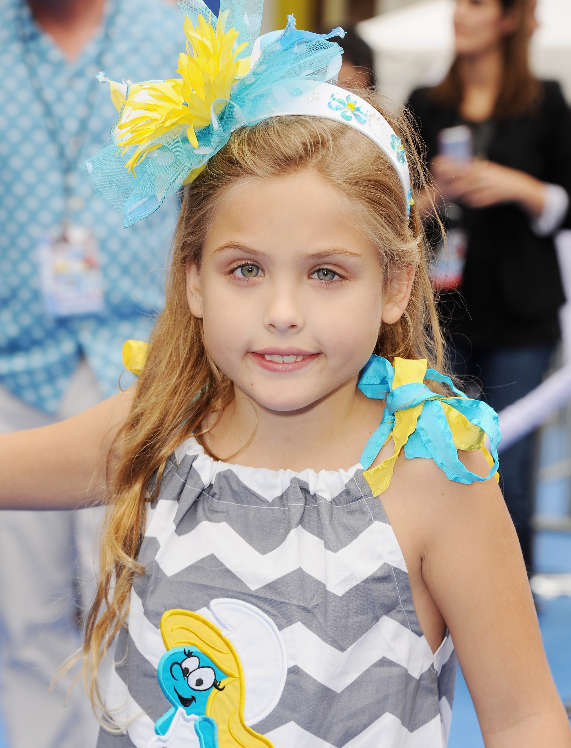 A vibrant oversized headband bursting with blue and yellow tulle and floral accents crowned 6-year-old Dannielynn Birkhead's long, flowing blonde hair at the Los Angeles premiere of "Smurfs 2" on July 28, 2013. Stepping onto the carpet at the Regency Village Theatre in Westwood, California, she radiated joyful confidence in a playful grey-and-white chevron dress featuring cheerful shoulder bows and a Smurf graphic, her bright eyes and warm smile highlighting her growing poise.
