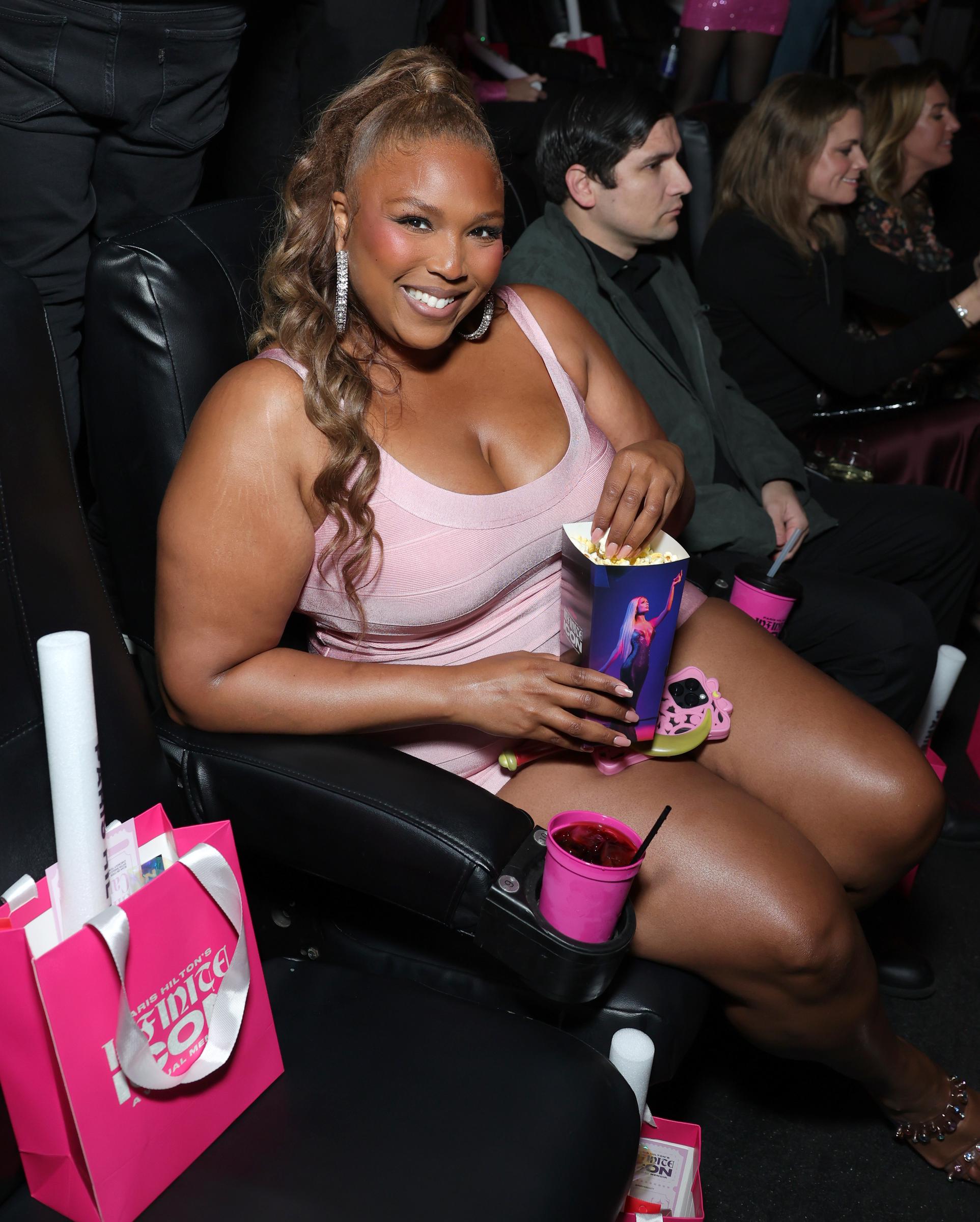 Captured mid-laugh in a seated shot, she holds a box of popcorn, and even in a relaxed position, her thighs appear slimmer and more toned. Her collarbone is visible, and her arms rest naturally with no excess fullness.
