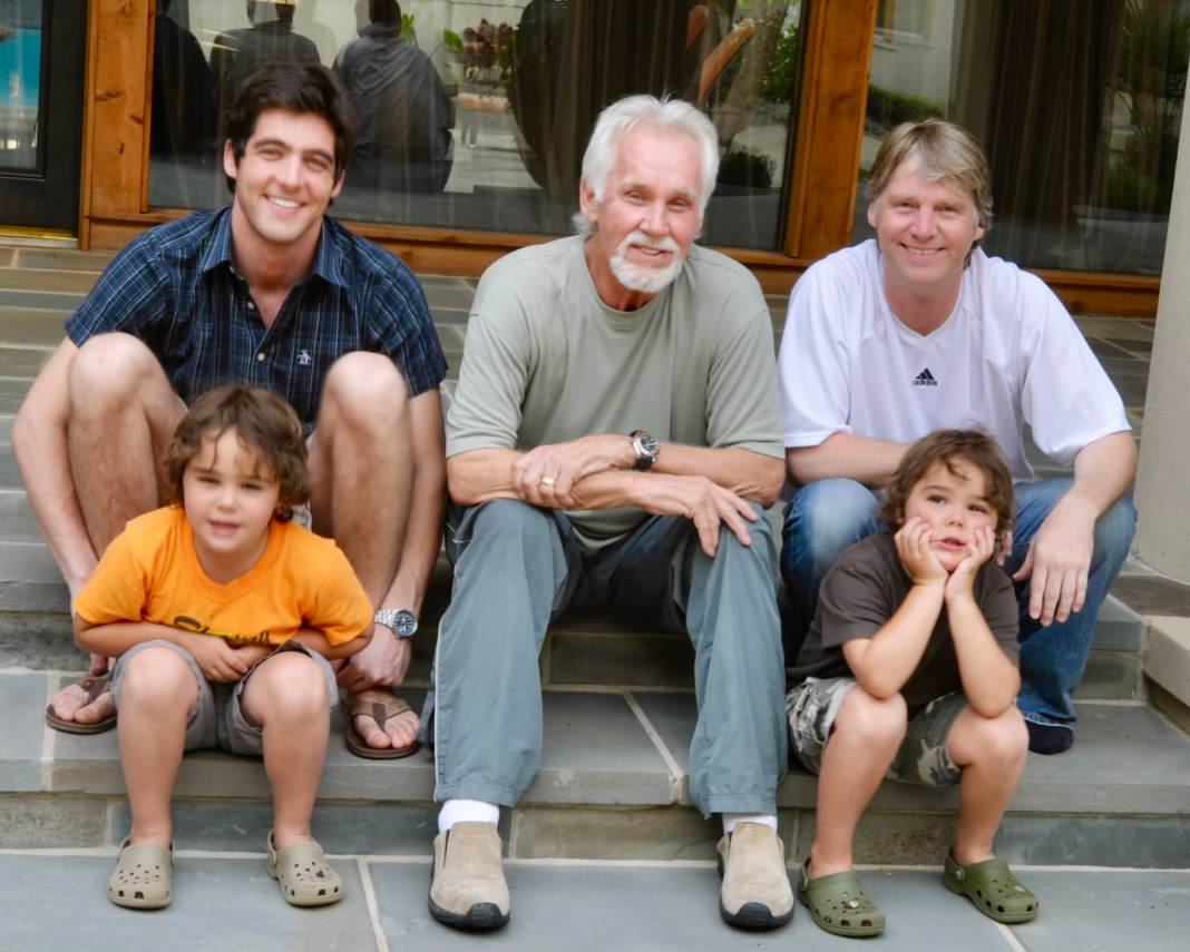 This childhood photo shows Justin and Jordan Rogers sitting with their father, Kenny Rogers, and other family members, capturing the country legend surrounded by his sons during the years when he was balancing the joys and concerns of later-in-life fatherhood. | Source: Facebook/Kenny Rogers