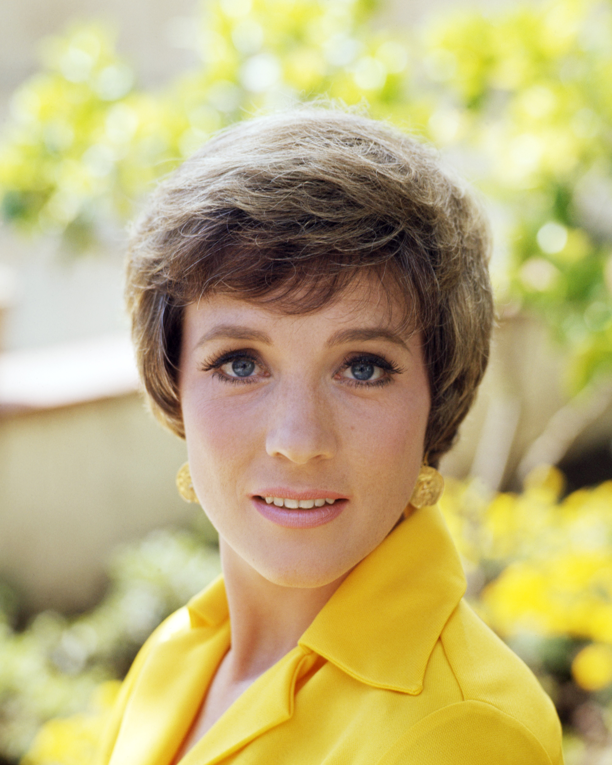 Julie Andrews radiates warmth in an outdoor color portrait taken circa 1965, smiling softly against a sunlit garden backdrop. The beloved English actress sports her trademark voluminous cropped pixie cut, pairing a collared bright yellow top with ornate gold button earrings that accent her striking blue eyes.