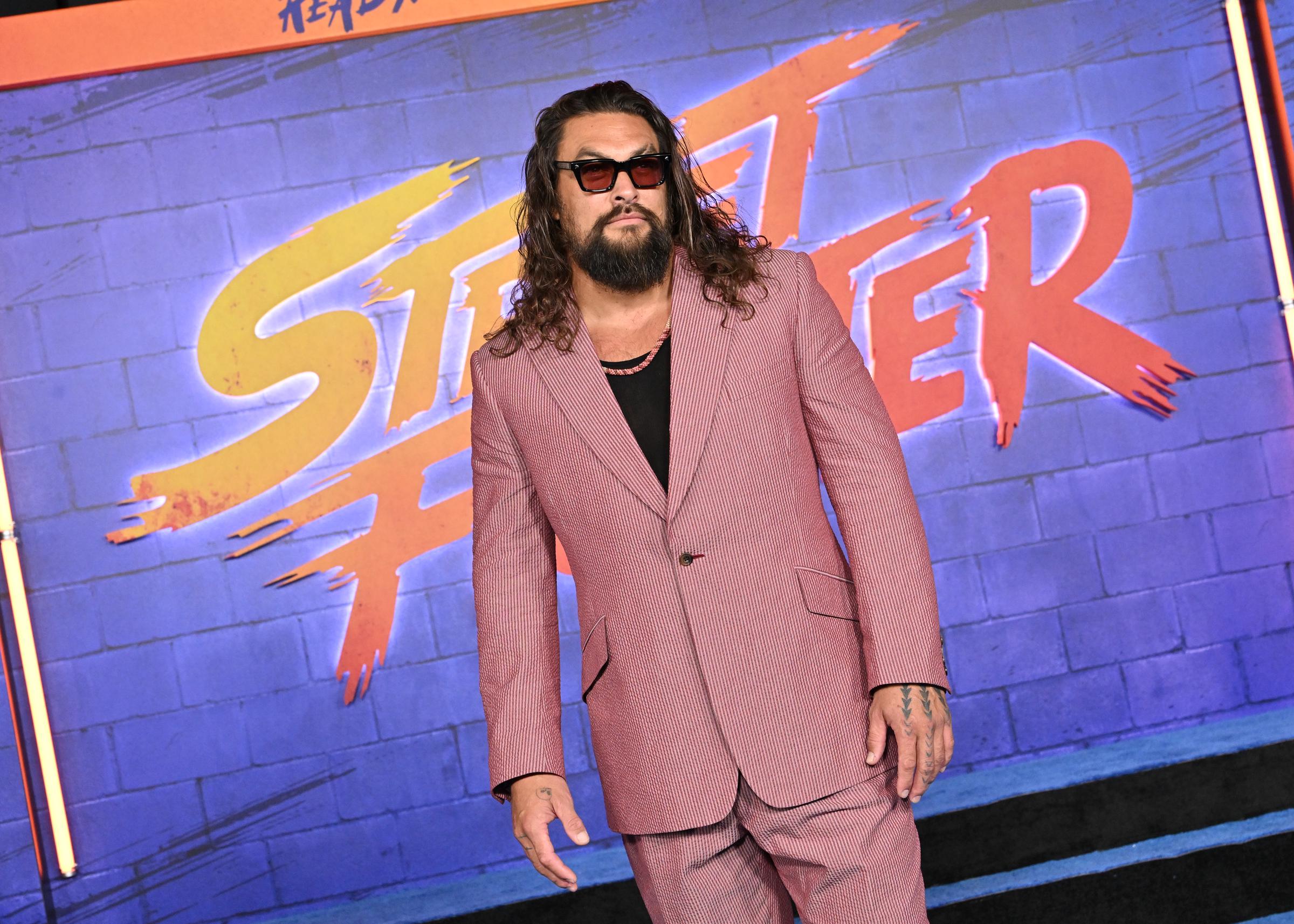 Captured in a three-quarter angle against the colorful neon title wall, the actor adopts a relaxed stance in his single-breasted suit. The black scoop-neck undershirt complements the warm red-and-white micro-patterned tailoring, while subtle hand tattoos peek out as his arms hang naturally at his sides. | Source: Getty Images