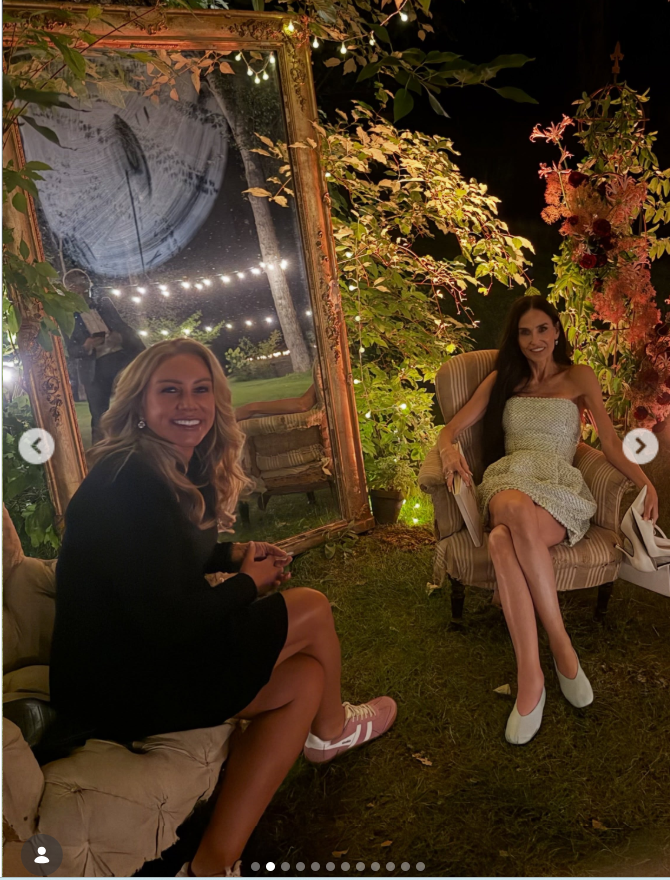 Demi Moore and Heather Parry are pictured relaxing outside during Tallulah Willis and Justin Acee’s wedding celebration. Demi wore her sleek, waist-length hair in a center part with diamond drop earrings and smoky eye makeup, while her short textured dress featured a woven pattern in silver and white. | Source: Instagram/heather_parry