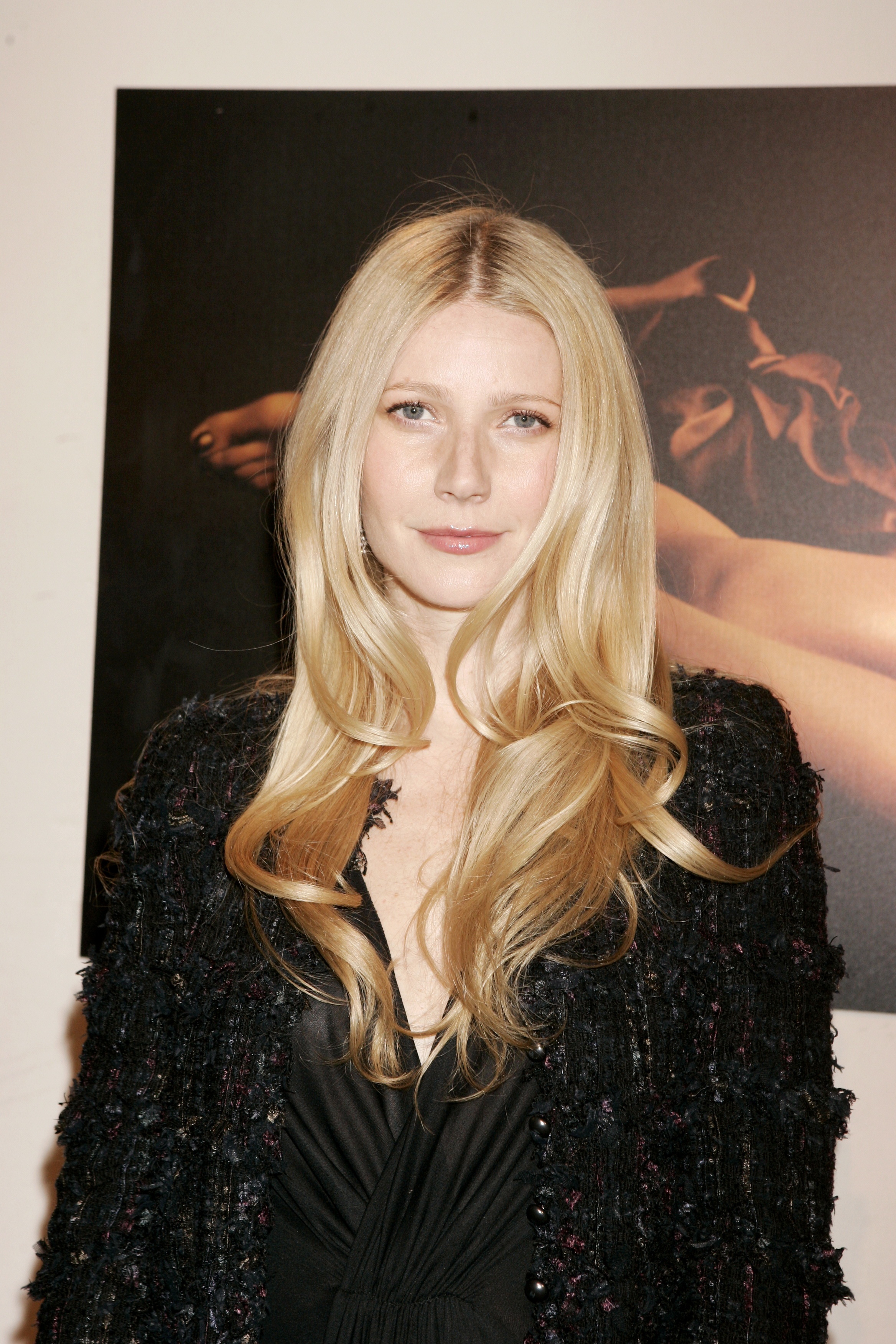 Gwyneth Paltrow attends the Tom Ford Estée Lauder Collection event in 2005, wearing a textured black jacket layered over a deep V-neck dress. Her long blonde hair is styled in loose, voluminous waves with a center part, while her natural makeup, highlighted by a soft pink gloss, gives her a luminous and elegant appearance.
