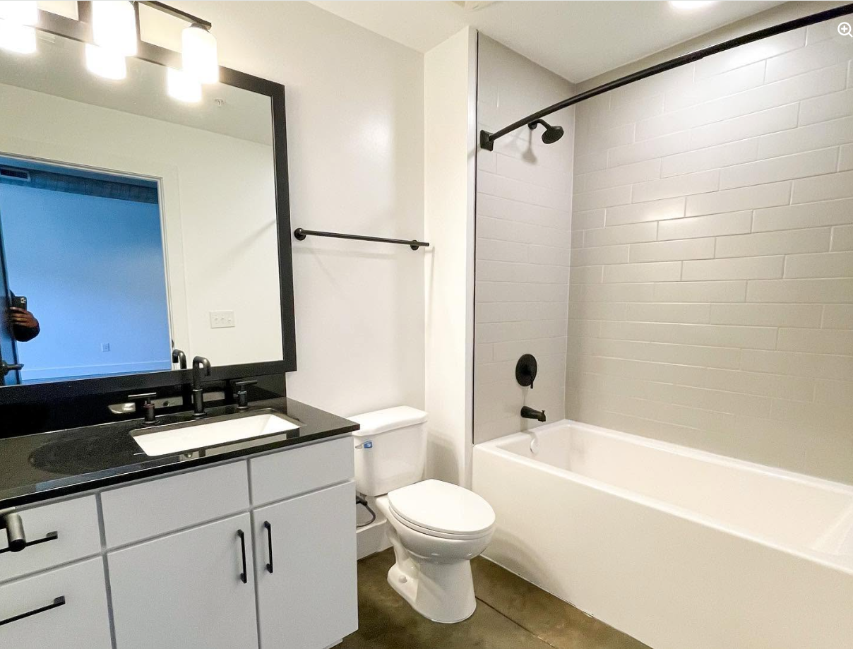 A bright bathroom features a white vanity, black fixtures and a subway-tile tub surround, continuing the simple, contemporary finishes seen throughout the loft. | Source: Facebook/Judson Mills Loft