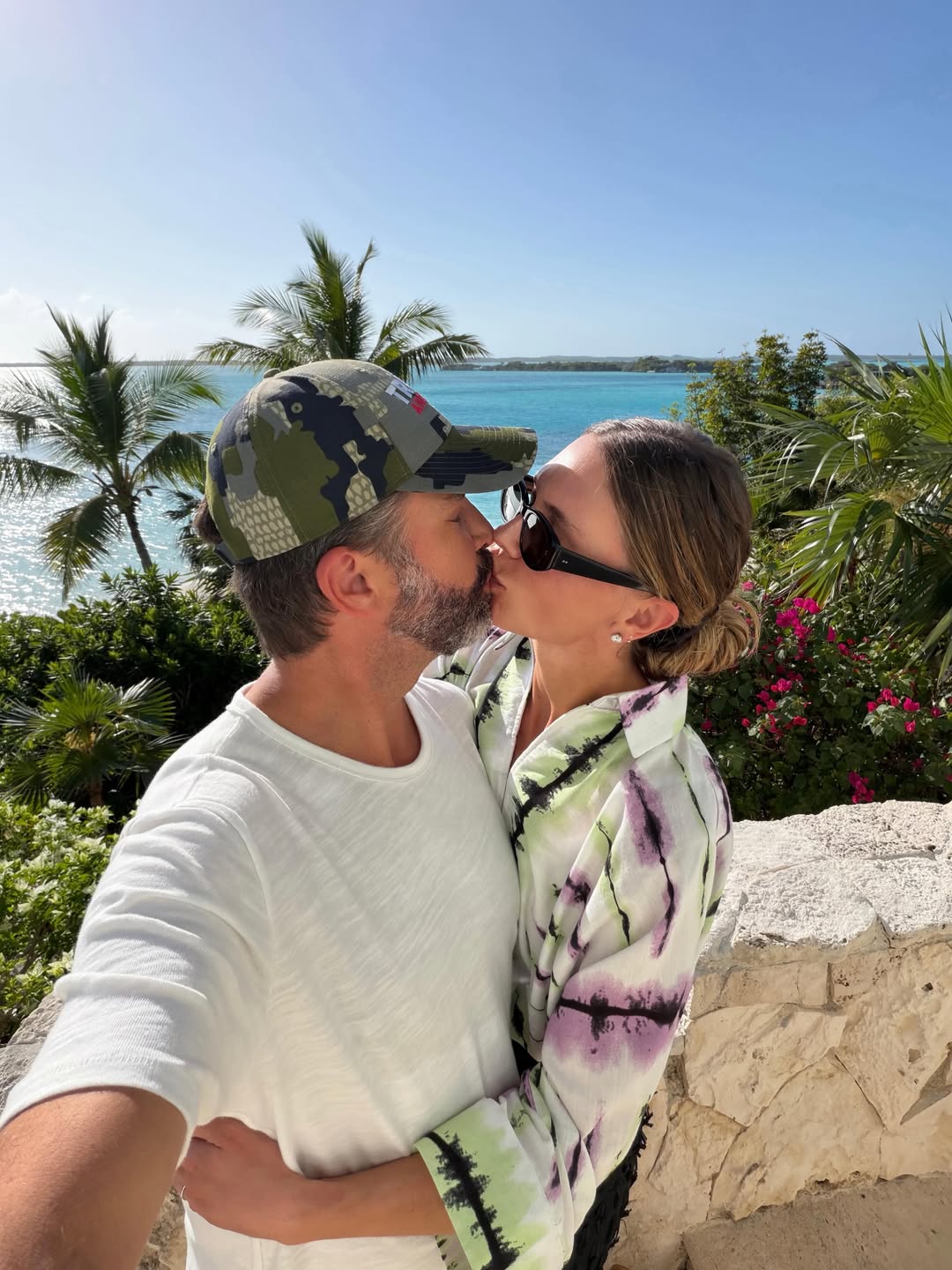 Donald Trump Jr. and Bettina Anderson share a kiss while posing for a selfie, with turquoise Bahamian waters and swaying palm trees stretching behind them. | Source: Instagram/bettinatrump