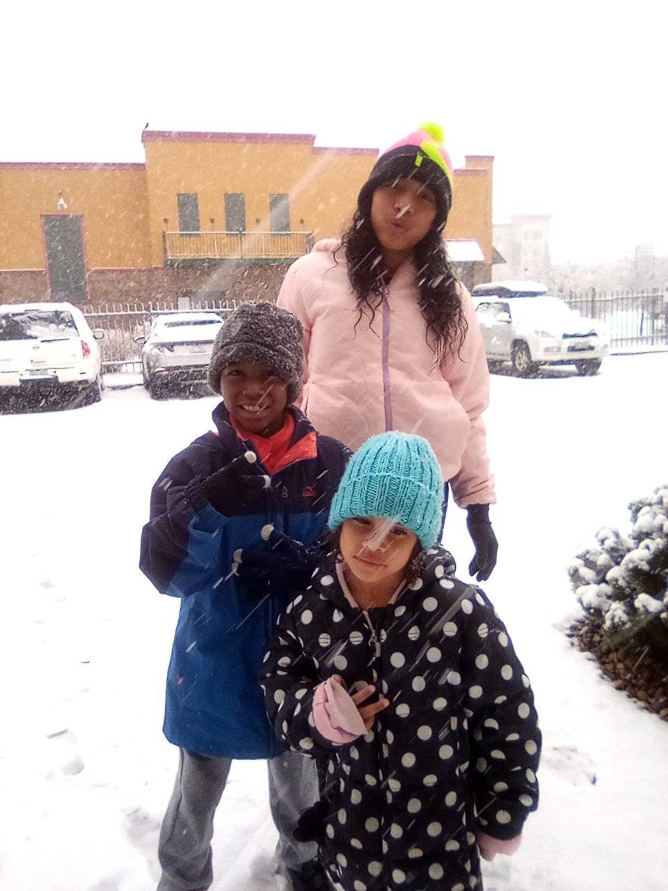 Eddimar Paola Hernández Chacón bundled up with her siblings during a snowy day outdoors. Eddimar stood in the foreground wearing a bright teal knit beanie and a black-and-white polka dot winter coat, posing alongside her brother in a blue-and-black hooded winter jacket and their older sister standing behind them in a pale pink puffer jacket as snow fell around them. | Source: Facebook/Morena Esa