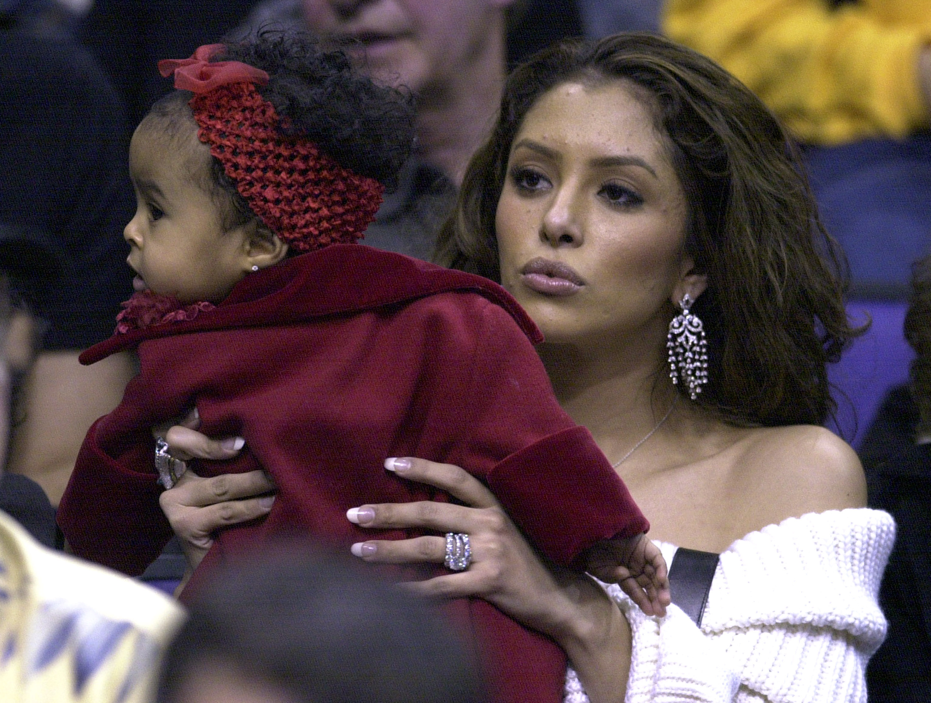 Vanessa Bryant cradles her young daughter, Natalia Diamante Bryant, as they watch the Los Angeles Lakers face the Houston Rockets at Staples Center, with Natalia dressed warmly in a red outfit and matching hair accessory.
