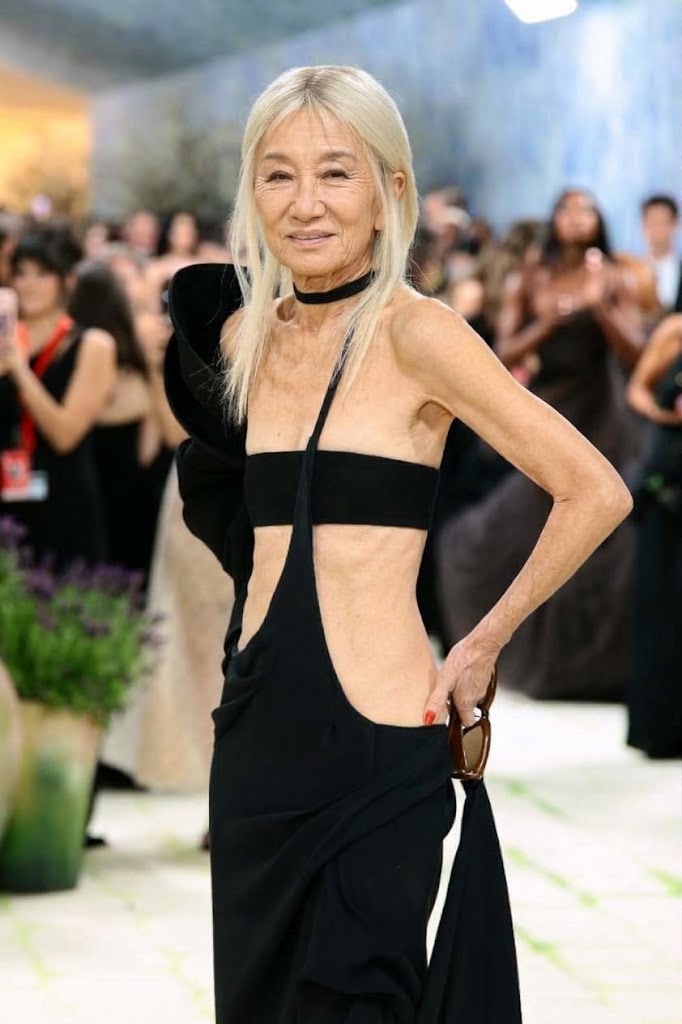 Drawing inspiration from the 2024 Met Gala green-and-white carpet, this AI creation reimagines Vera Wang's daring high-fashion appearance with visible, authentic signs of aging along her neck, décolletage, and midriff. The concept retains the designer's striking platinum-blonde hair, black neck choker, and cutout gown with an asymmetrical shoulder drape, presenting an untouched, natural facial structure with softened mature features. | Source: Gemini