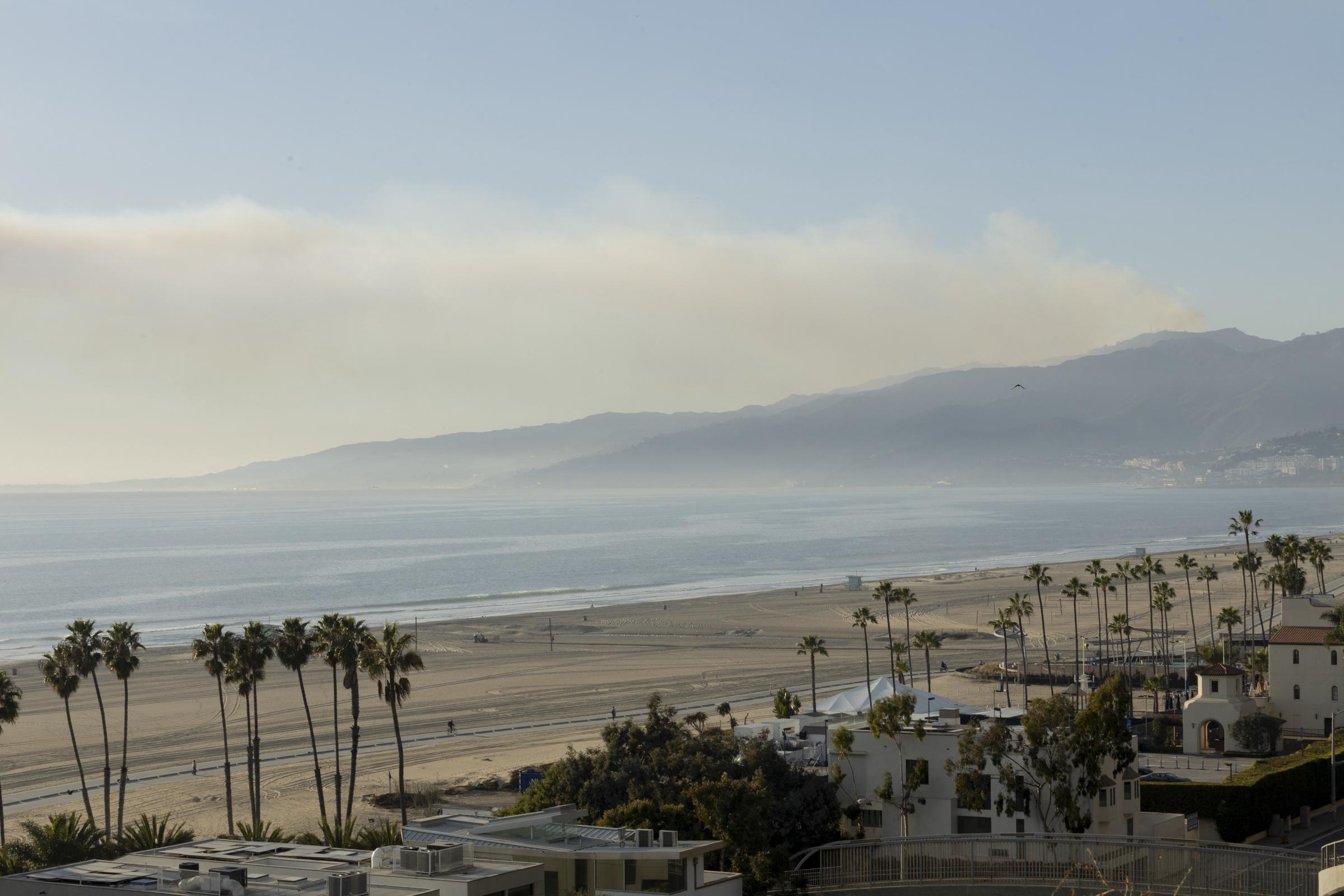 A wide elevated view captured Santa Monica Beach in Southern California on January 10, 2025, looking out over rows of tall palm trees, coastal structures, and the broad sandy shore toward the ocean. In the background, rolling coastal mountains stretched beneath a pale sky marked by a soft band of haze and smoke hanging over the horizon. | Source: Getty Images