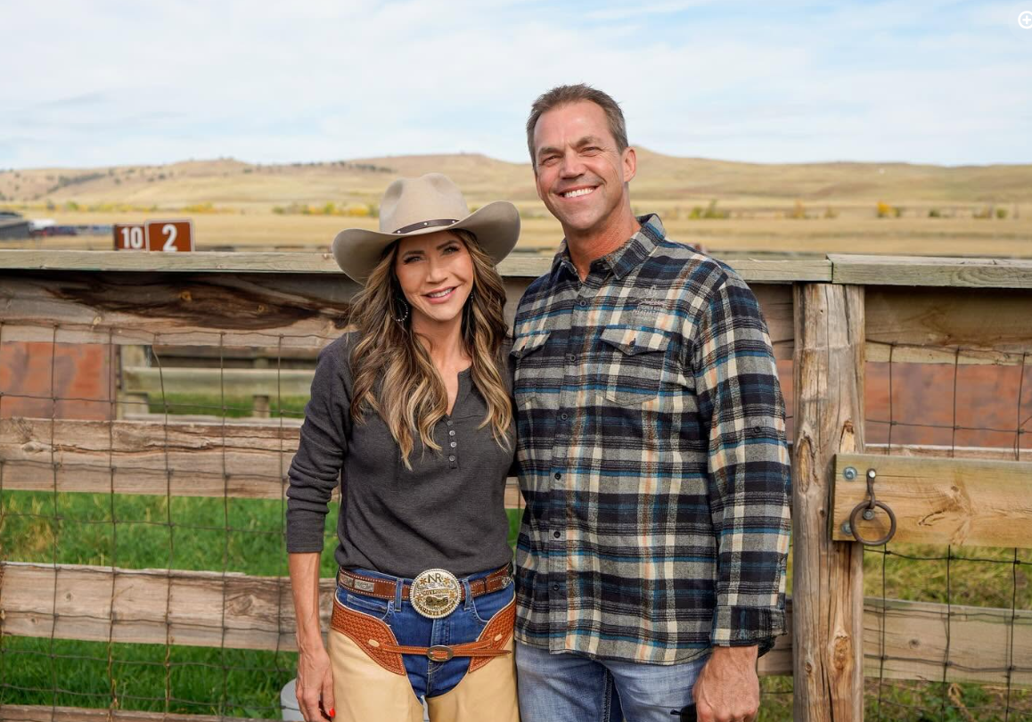 Kristi and Bryon Noem are pictured together in a ranch setting, with wooden fencing and open land behind them. The scene reflects the rural surroundings that appear throughout photographs from their life in South Dakota. | Source: Facebook/kristi.noem