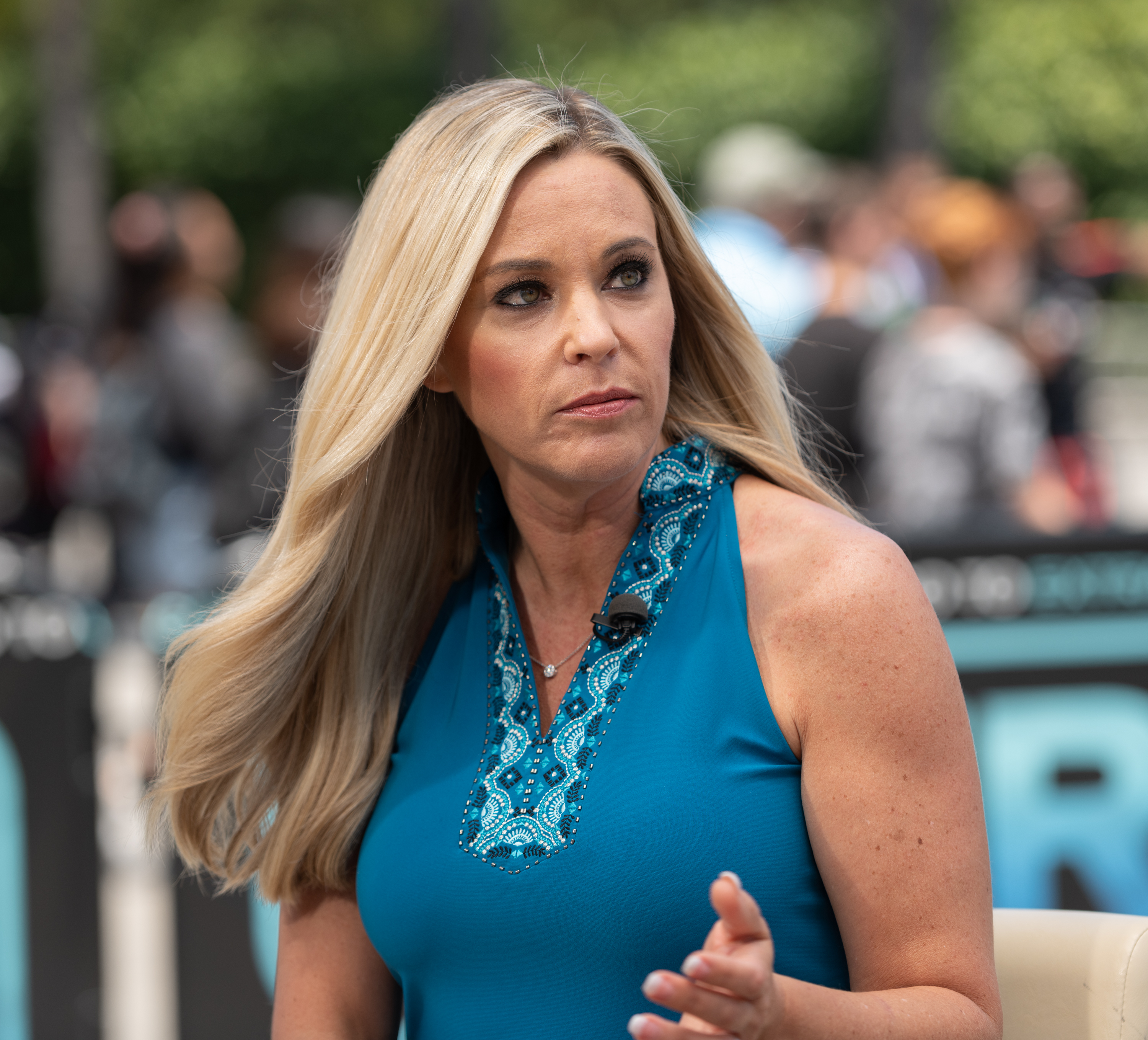 Speaking during an "Extra" interview outdoors, Kate Gosselin wears a bold teal halter top with jeweled detail. Her long blonde hair frames a focused expression as she gestures mid-conversation with confidence.