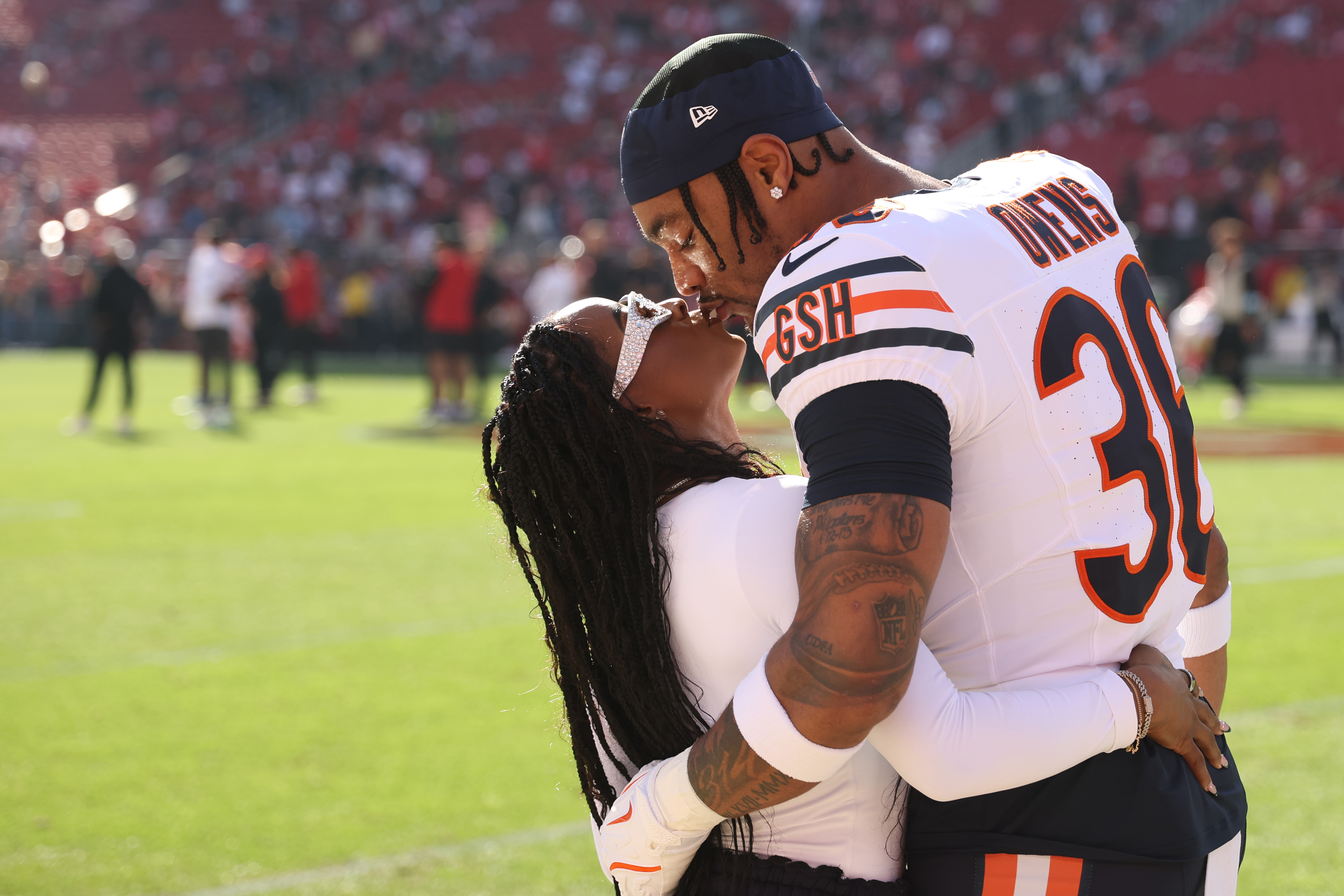 Simone Biles shares a kiss with husband Jonathan Owens on the field at Levi's Stadium on December 8, 2024. Owens, suited up in his Chicago Bears uniform, holds Biles close as she leans back with her arms around him, wearing stylish sunglasses and a long-sleeve white top.