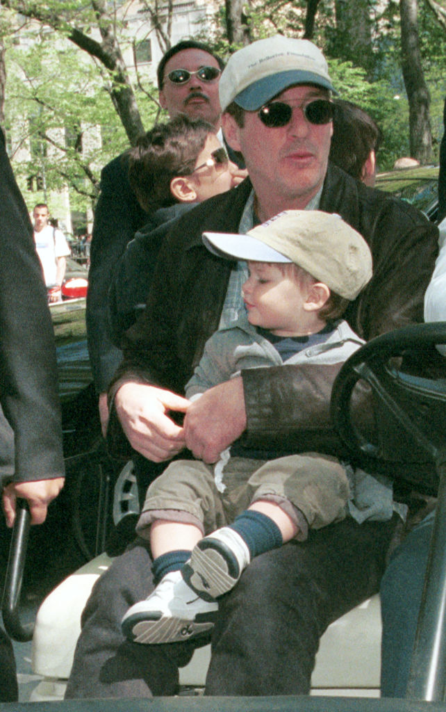 Wearing matching caps, Richard Gere holds Homer Gere close while arriving at the "Kids for Kids" Carnival in Central Park, New York City. The two sit together, calmly taking in the event around them.