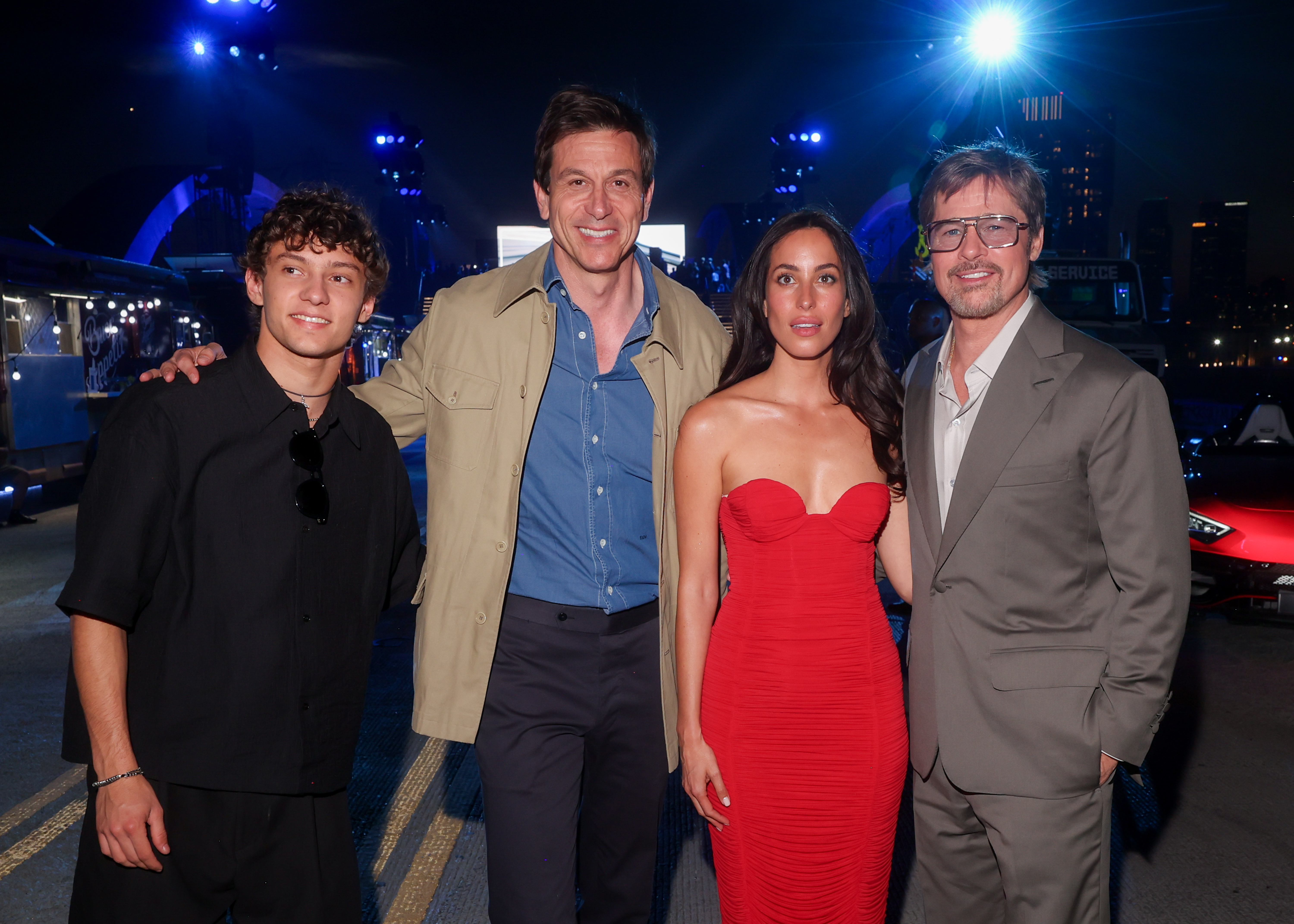 Andrea Kimi Antonelli, Toto Wolff, Ines de Ramon, and Brad Pitt gather for a group photo surrounded by bright lights, luxury cars, and guests at the premiere.