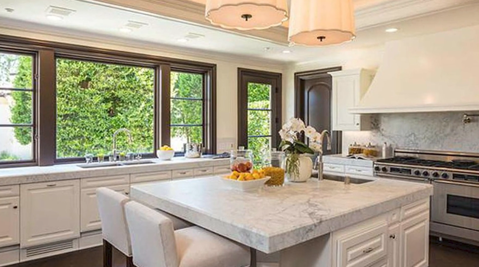 The expansive gourmet kitchen centers on an oversized marble-topped island with casual counter seating, surrounded by white raised-panel cabinetry, a commercial-grade stainless steel range with a custom hood, and broad dark-trimmed windows looking out to private greenery. | Source: YouTube/Taste of Country