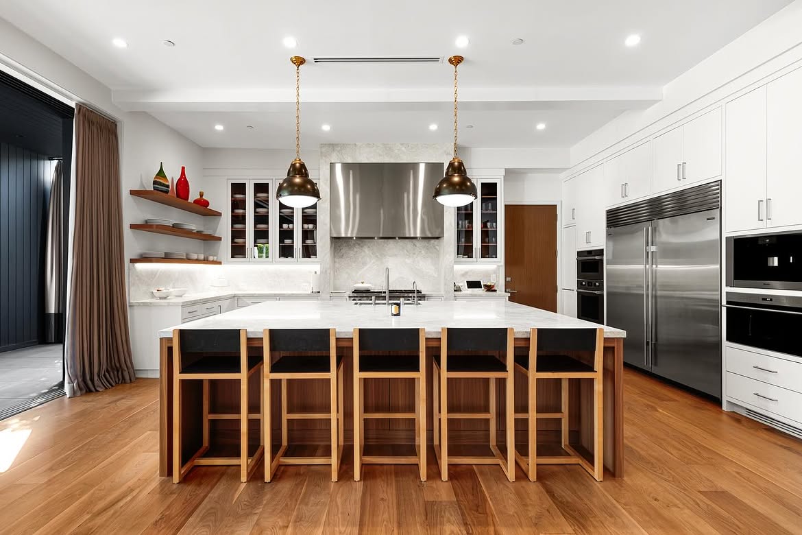 The broad island dominates the kitchen, providing seating for five beneath two dark pendant lights. Stainless-steel appliances, including an oversized refrigerator and range hood, break up the extensive white cabinetry. | Source: Instagram/theshevins