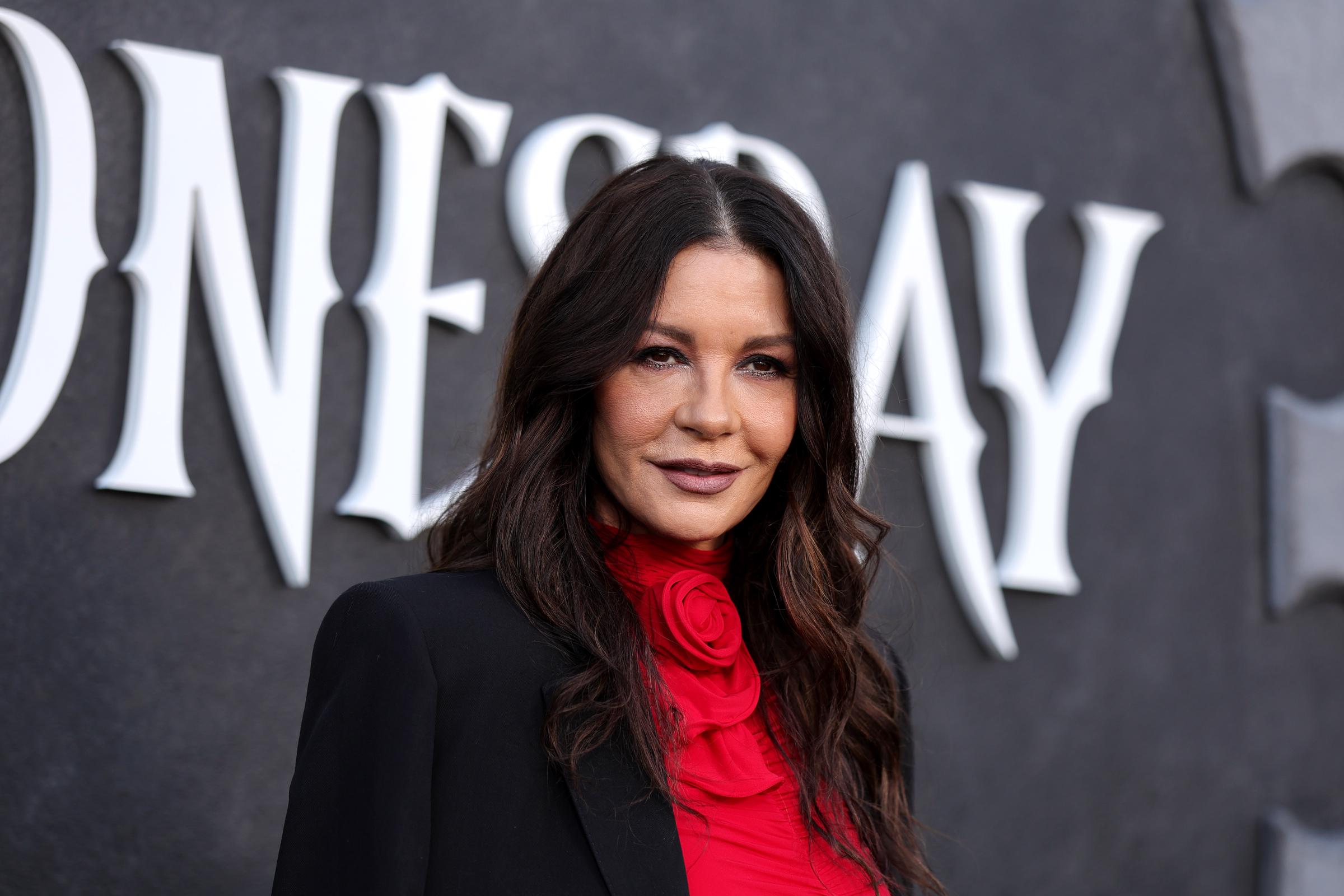 Catherine Zeta-Jones poses against the themed backdrop during Netflix's "Wednesday" Emmys FYSEE event at Hollywood Forever Cemetery on May 22, 2026, in Los Angeles, California. The actress wears a tailored black blazer layered over a vibrant crimson Magda Butrym top featuring an ornate rosette neck detail, styling her brunette hair in relaxed middle-parted waves with smoky eye makeup and a soft mauve lip.