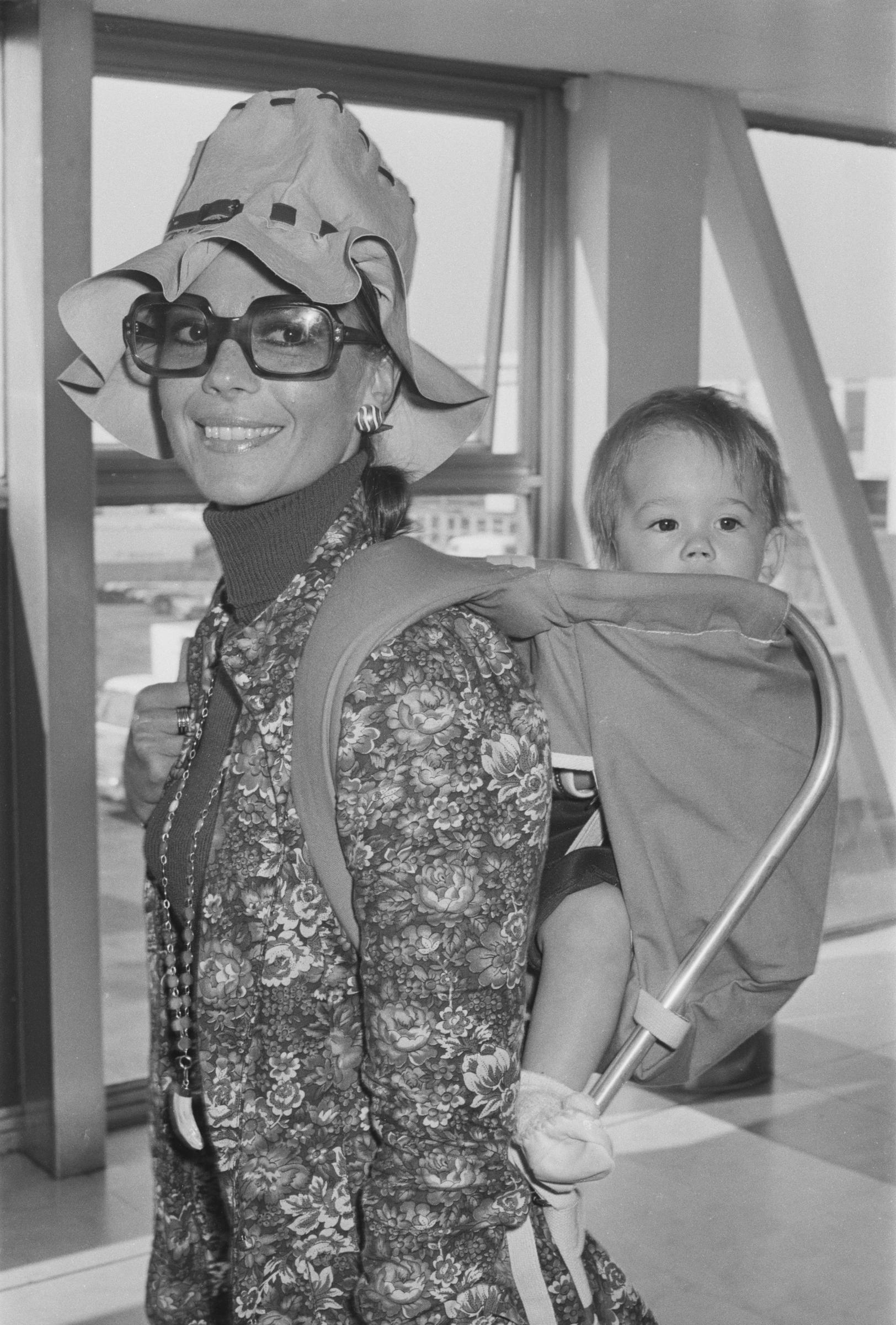 Natalie Wood smiles at the camera while carrying daughter Natasha Gregson Wagner in a baby carrier on her back. Taken in the UK on September 21, 1971, Natalie wears oversized glasses, a wide-brimmed hat, and a floral-patterned coat. Natasha peeks over her shoulder, bundled in a hooded outfit.