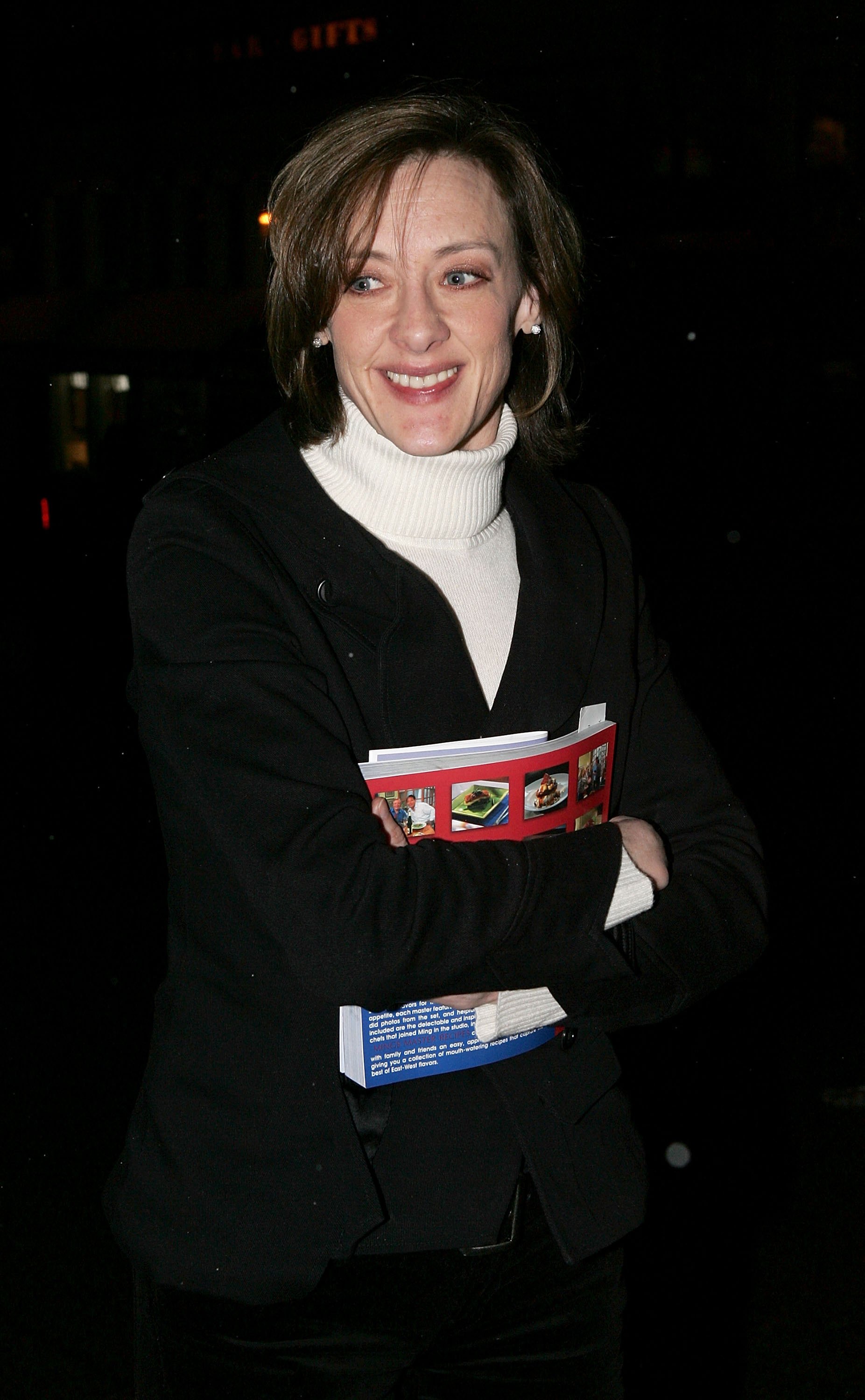 Joan Cusack smiles warmly while clutching a colorful book against a crisp white turtleneck and dark blazer. Against the dark nighttime backdrop, her relaxed expression gives the candid moment a cozy feel.