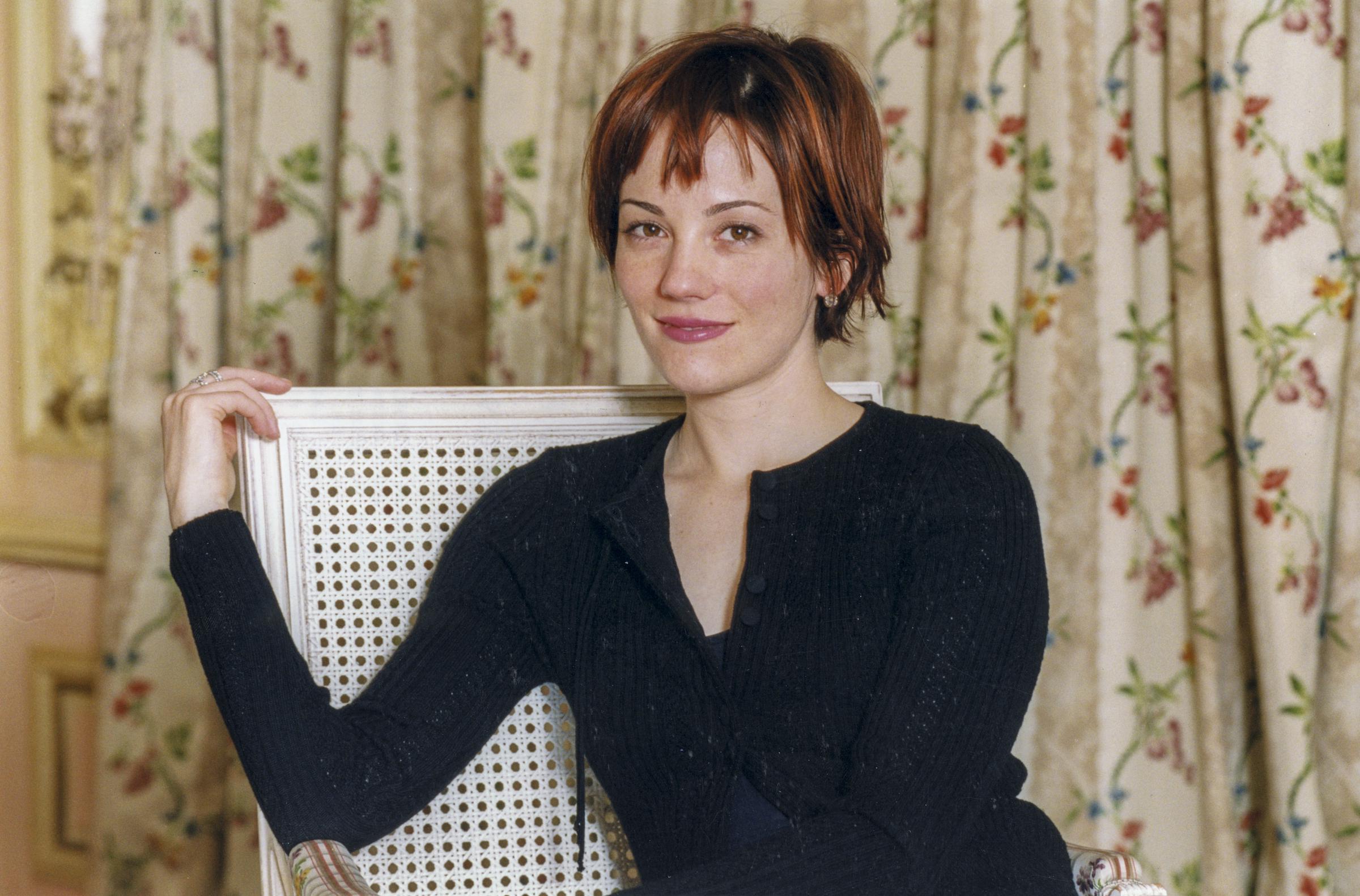 Natasha Gregson Wagner poses for a portrait at the Festival du Film Policier de Cognac in France, March 1999. She wears a black ribbed cardigan, seated on a white cane-back chair in front of floral curtains, with a soft smile and short auburn hair.
