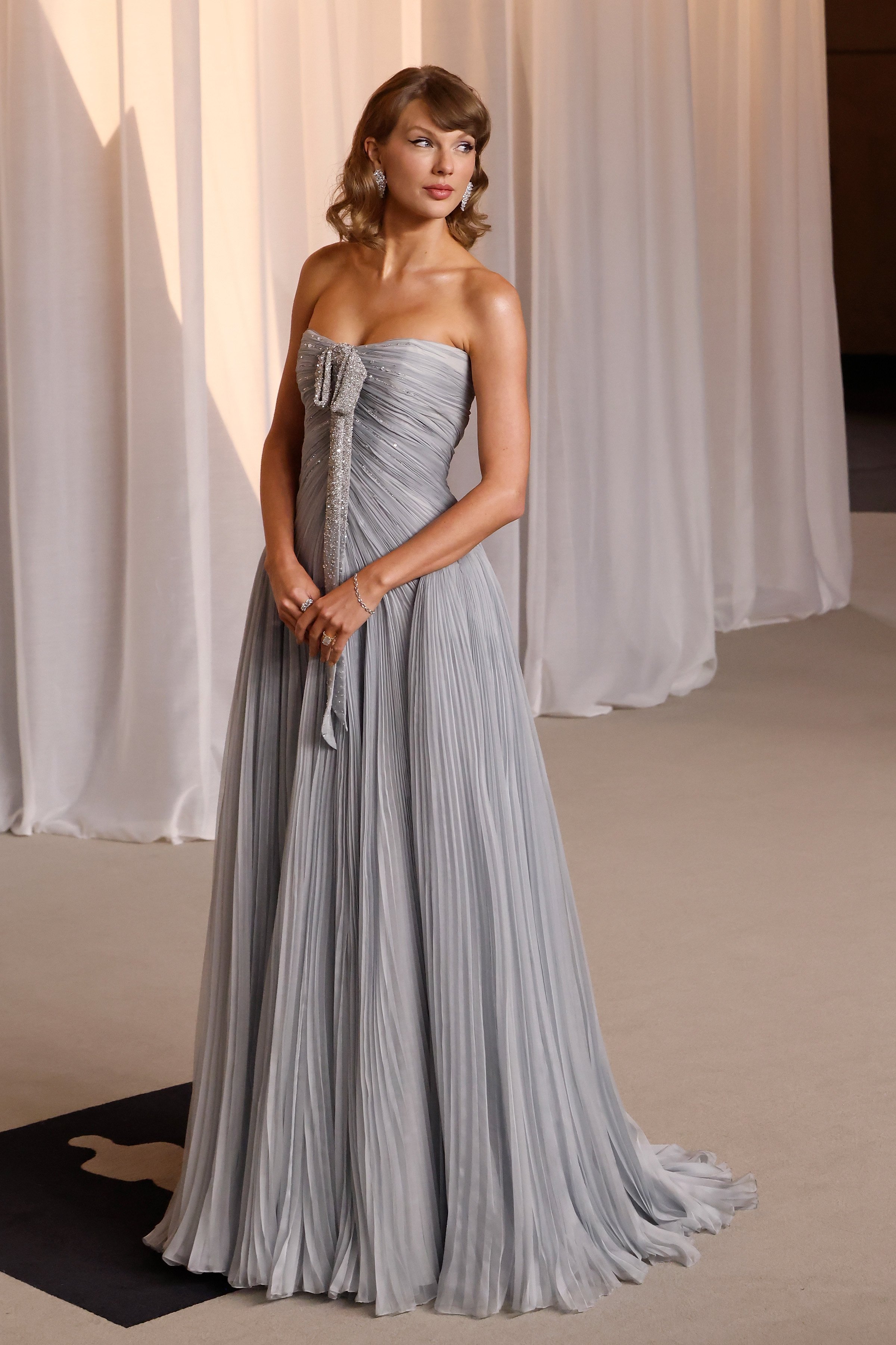 Taylor Swift radiates ethereal elegance in a strapless dove-gray gown accented by delicate pleating throughout the bodice and sweeping skirt. A shimmering, crystal-embellished bow detail cascades gracefully down the front, complemented by sparkling diamond drop earrings and soft vintage waves. Ready to take the stage as the evening's featured musical performer, the Grammy winner delivers pure timeless glamour.