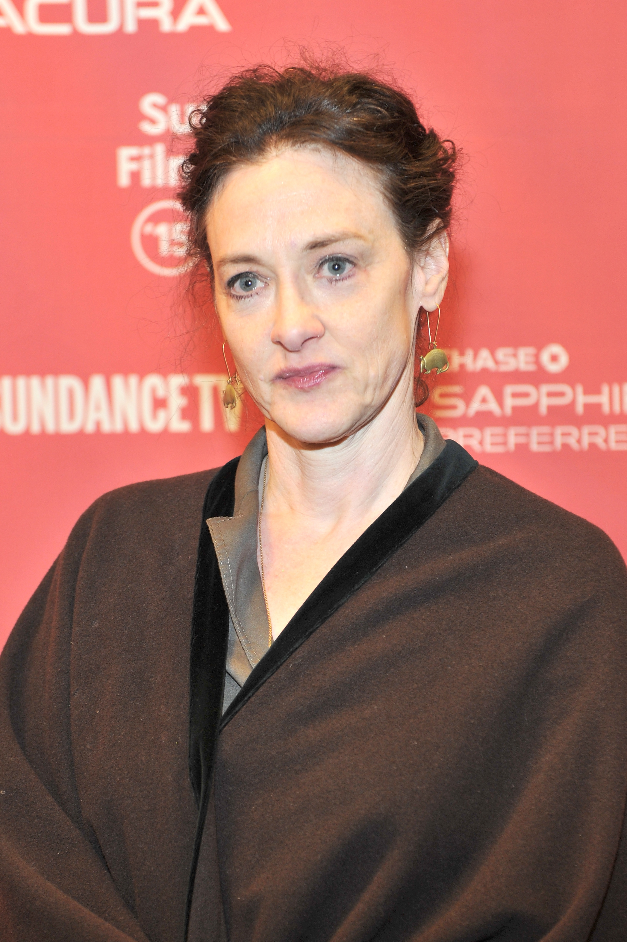 Joan Cusack appears poised and understated in a dark layered outfit with her hair swept back. Set against a bright red backdrop, her thoughtful expression gives the portrait a quiet intensity.