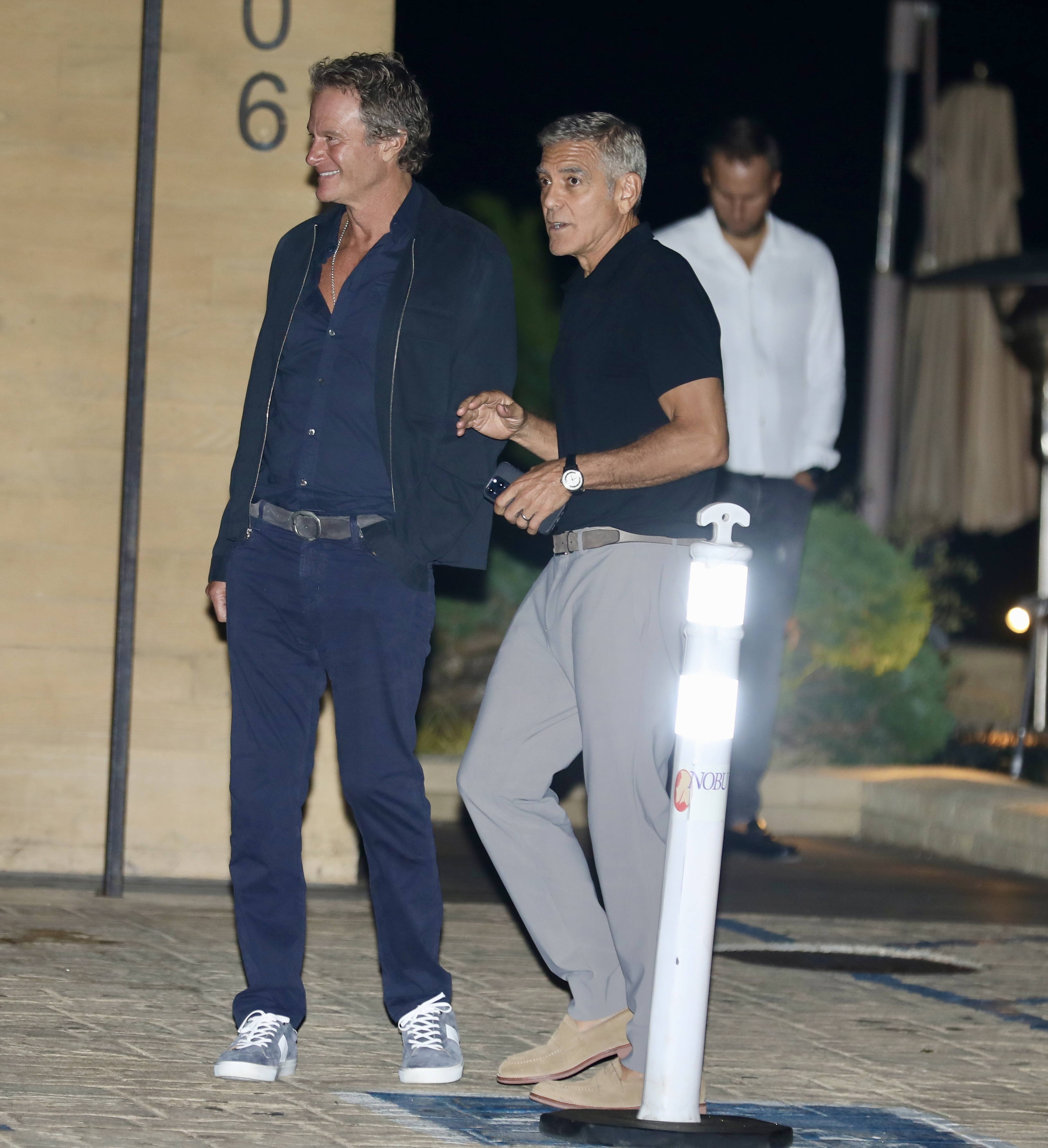 Close pals Rande Gerber and George Clooney kept it smart-casual for a dinner outing at Nobu on September 14, 2024, in Malibu, California. Strolling side by side into the evening, Rande rocked a navy jacket over an open-collared navy shirt, trousers, and light blue sneakers, while Clooney sported a black polo paired with light gray chinos, tan suede shoes, and his phone in hand. | Source: Getty Images