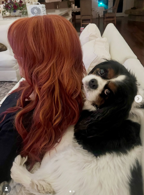 A small black-and-white dog rests against Wynonna as she sits on a white sofa. The photograph offers a quieter domestic moment among the cooking scenes and outdoor farm activities she has shared. | Source: Instagram/wynonnajudd
