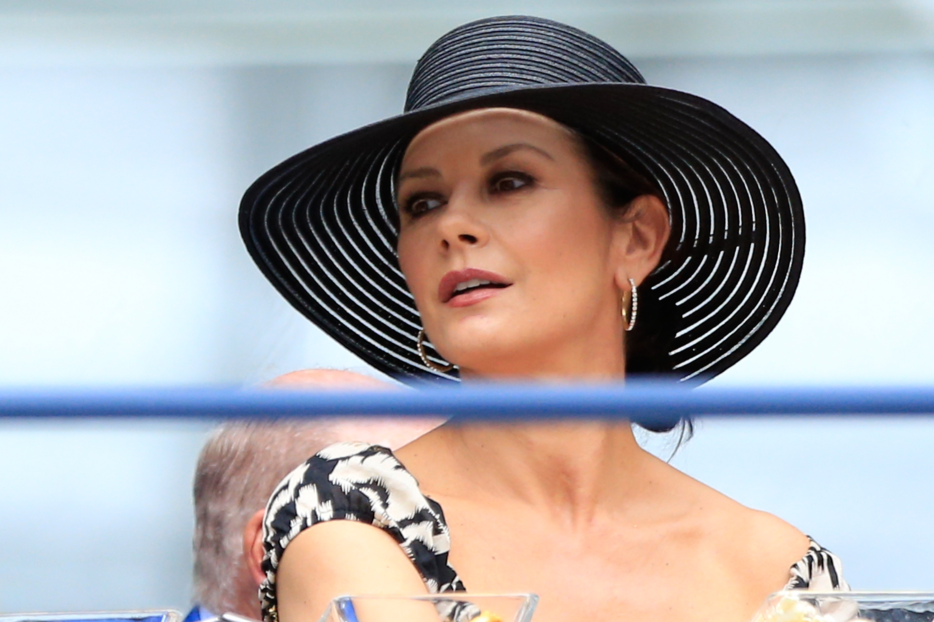 Catherine Zeta-Jones watches the Women's Singles Final match between Roberta Vinci and Flavia Pennetta from the stands during Day Thirteen of the 2015 US Open at the USTA Billie Jean King National Tennis Center on September 12, 2015, in Flushing, Queens, New York City. She looks chic in a wide-brimmed black woven sun hat, hoop earrings, and an off-the-shoulder black-and-white patterned dress, gazing attentively out toward the court.