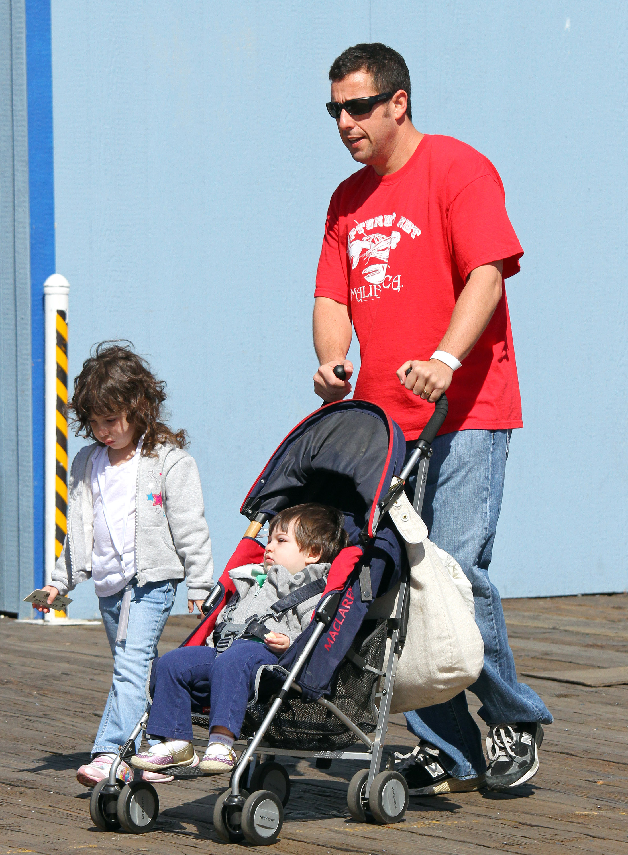 Adam Sandler strolls through Santa Monica, pushing a stroller while Sadie Madison Sandler walks beside him, absorbed in her own little world. Dressed casually for the day, the scene captures a simple family outing — quiet, unguarded, and grounded in everyday life.