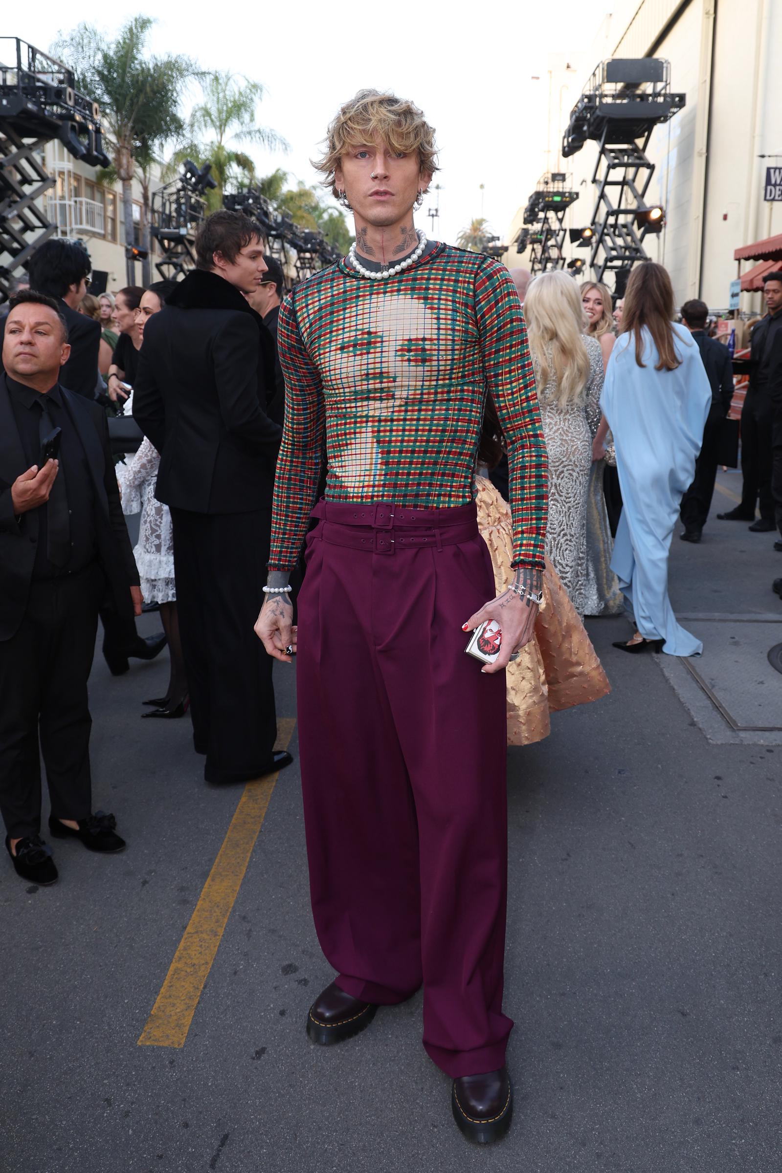Musician mgk makes a bold fashion statement at Vogue World: Hollywood 2025 at Paramount Studios in Los Angeles on October 26, 2025. The artist stands out on the outdoor carpet in a graphic, sheer plaid long-sleeve knit top styled with a pearl necklace, high-waisted belted plum-purple wide-leg trousers, and dark lug-sole shoes. | Source: Getty Images