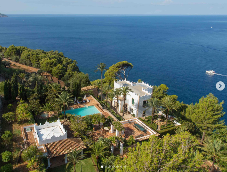 S'Estaca's clifftop setting puts the Mediterranean at the center of the estate, with the white main house, pool, terraces, and surrounding greenery spread above the water. The aerial view captures the dramatic coastal setting that first drew Douglas to the property in 1990. | Source: Instagram/robbreport