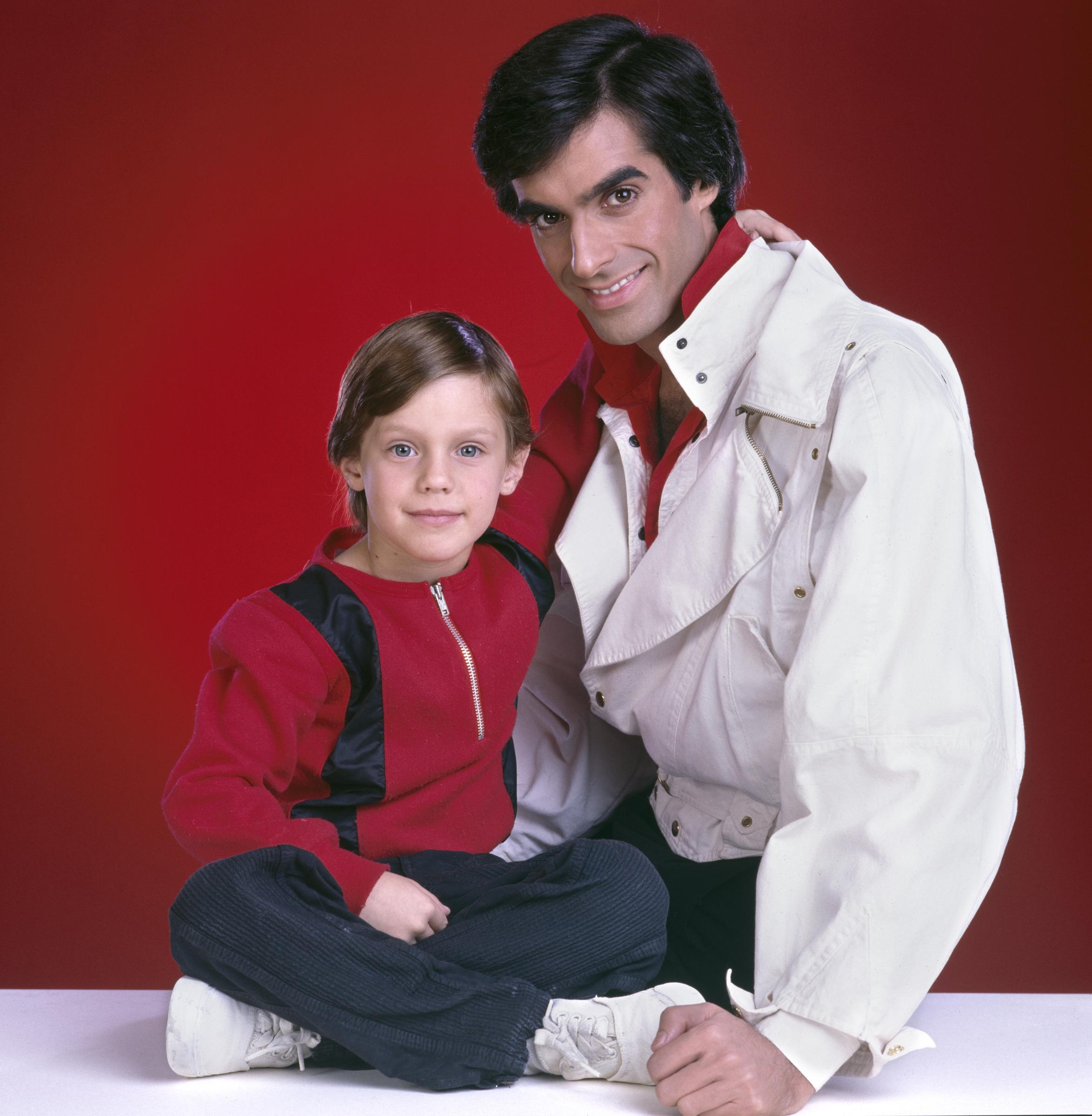 A young Omri Katz sits cross-legged with a neat, side-parted chestnut haircut, bright wide eyes, and a sweet smile while wearing a red-and-black zip-up sweatshirt with dark corduroy pants. He poses alongside illusionist David Copperfield for the television special "The Magic of David Copperfield VII" (also known as "The Magic of David Copperfield VII: Familiares"), originally broadcast on March 8, 1985.
