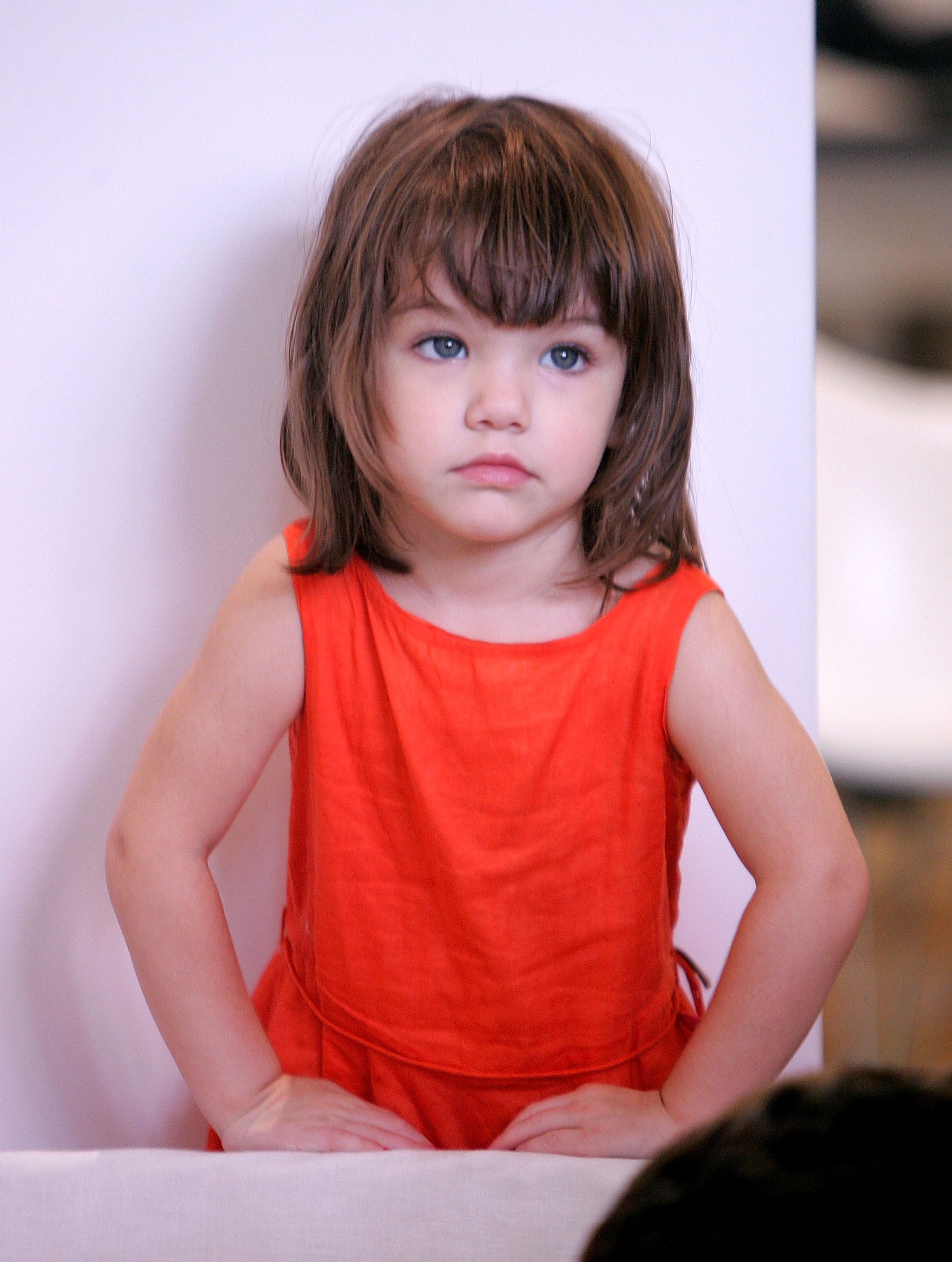 Suri Noelle was photographed in New York City's West Village on August 17, 2008, wearing a bright coral dress as she looked toward the camera with a thoughtful expression. Still a toddler at the time, she had already become one of Hollywood's most recognizable celebrity children, frequently drawing attention during public outings with her famous parents.