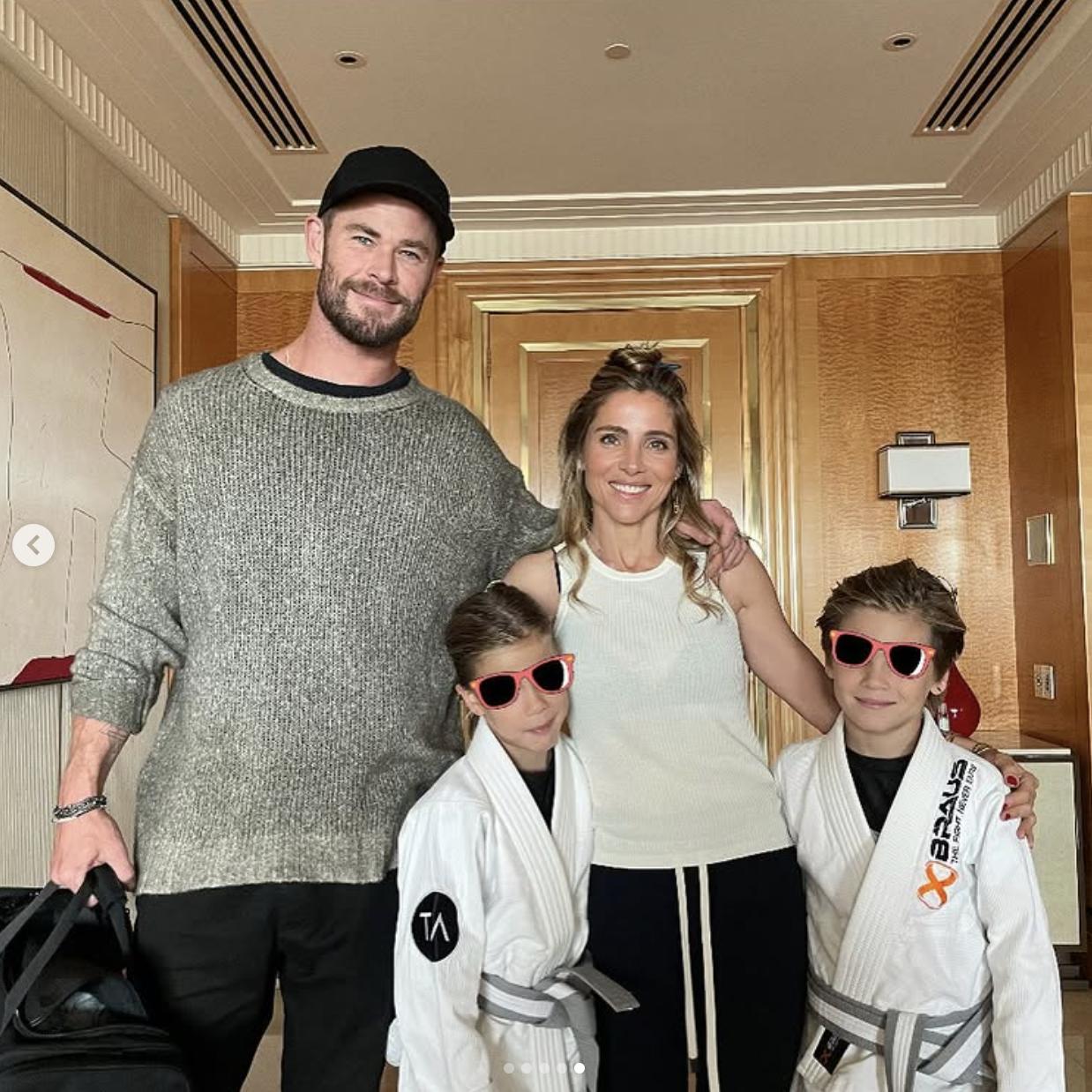 Chris Hemsworth and Elsa Pataky pose with their twin sons, Tristan and Sasha, following a successful jiu-jitsu competition in November 2022. The boys wear matching white gis and red sunglasses, each proudly displaying their belts after earning two gold medals. The family stands together inside what appears to be a hotel room, beaming after the achievement.