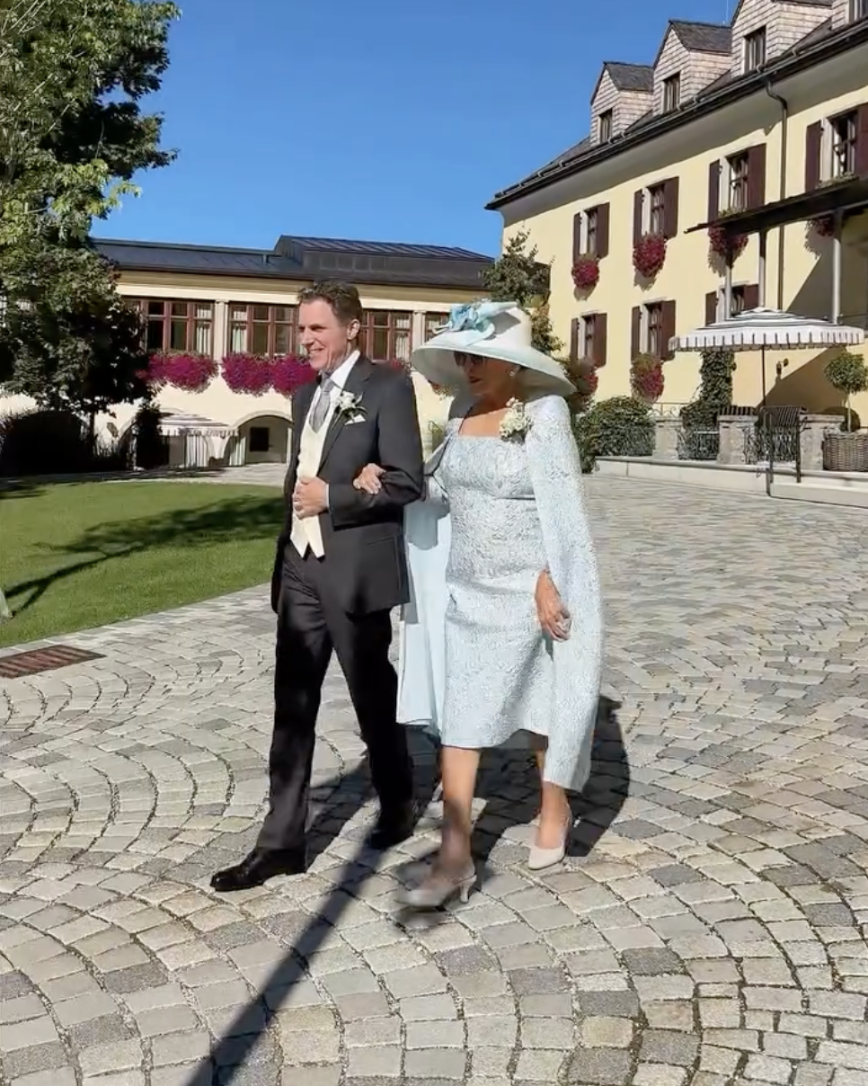 With elegance and poise, Joan Collins accompanies Alexander Newley before the ceremony. Her blue jacquard ensemble and matching hat shine against the crisp backdrop of the wedding venue. | Source: Instagram/sheelanewley