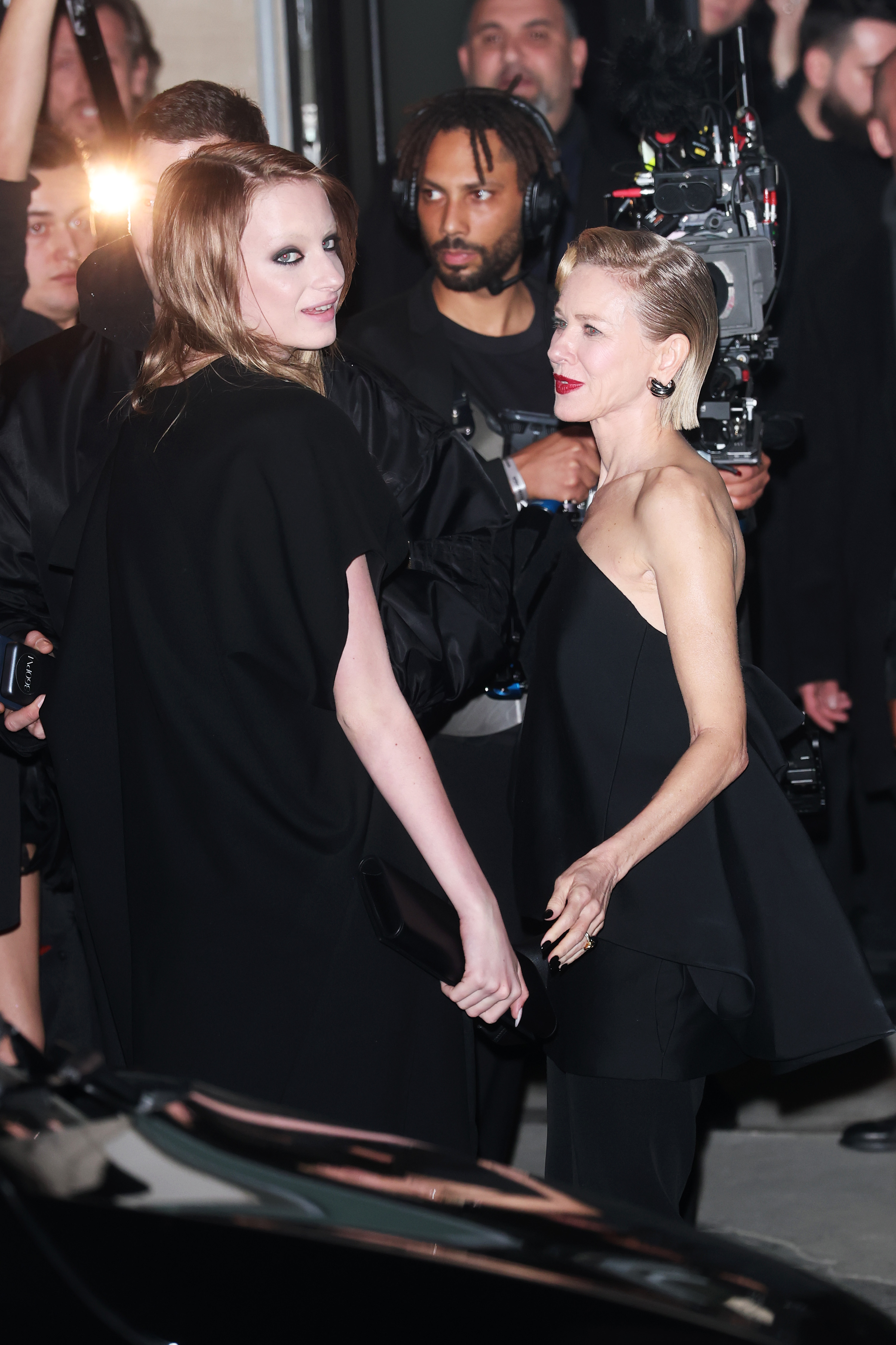 Kai Schreiber and Naomi Watts share a moment outside the Balenciaga show during Paris Fashion Week. Kai glances back toward the cameras while Watts speaks beside her. The mother-daughter duo move through a crowd of photographers and crew.
