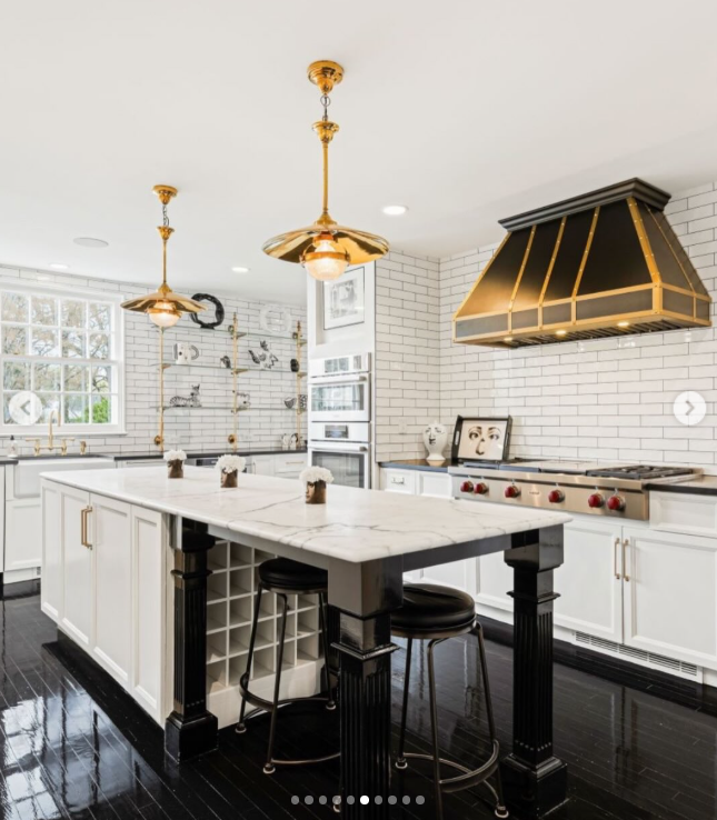 The kitchen takes a lighter approach with white cabinetry, subway tile, marble surfaces, and black flooring. Gold-toned fixtures and a dramatic range hood add warmth, while the large island provides seating and built-in wine storage. | Source: Instagram/rogerperrygroup