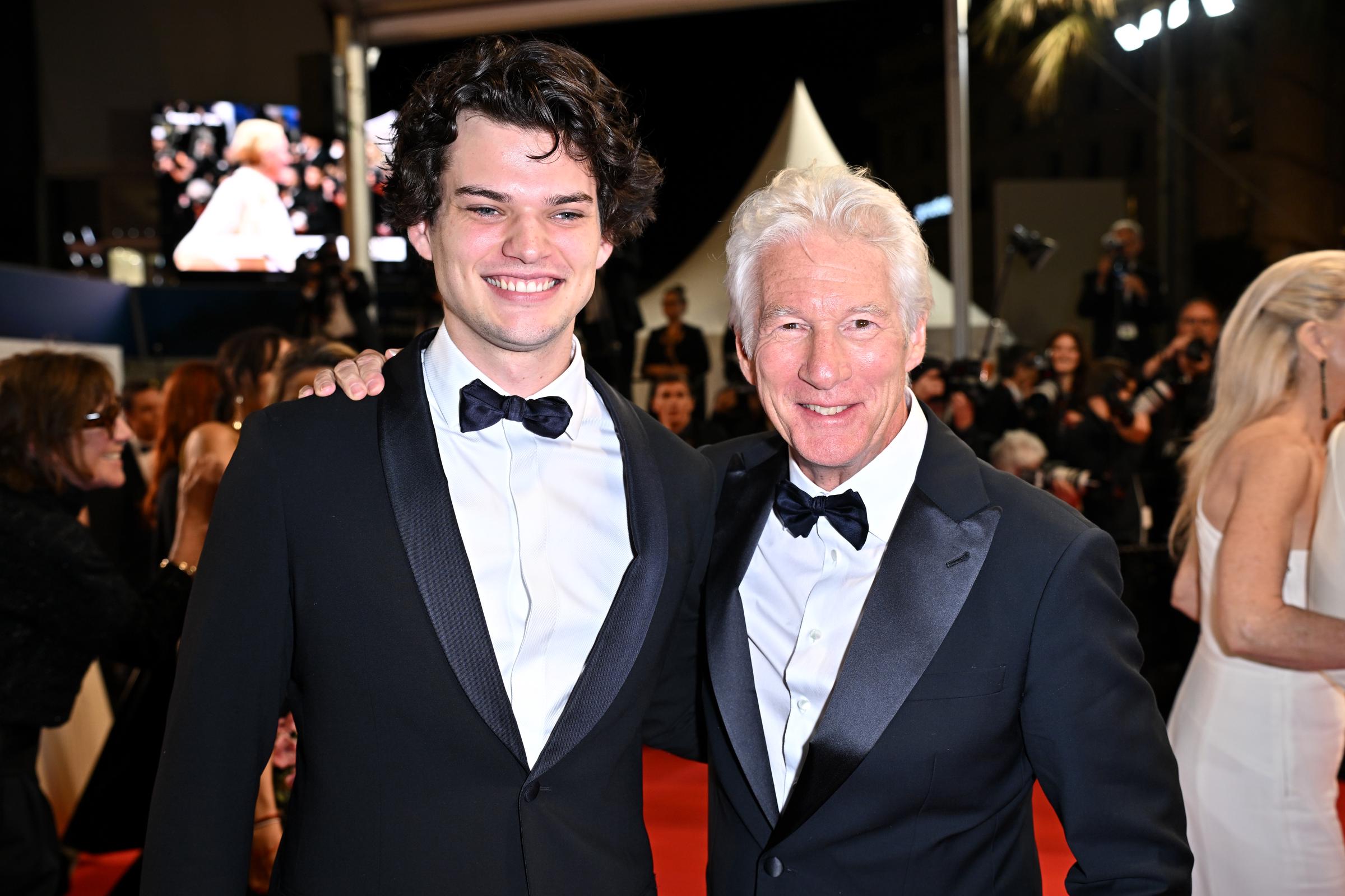 Homer and Richard Gere share a proud red carpet moment at the 77th Cannes Film Festival. Dressed in matching tuxedos, they smile side by side amid a swirl of flashing lights and elegant crowds at the "Oh, Canada" premiere.