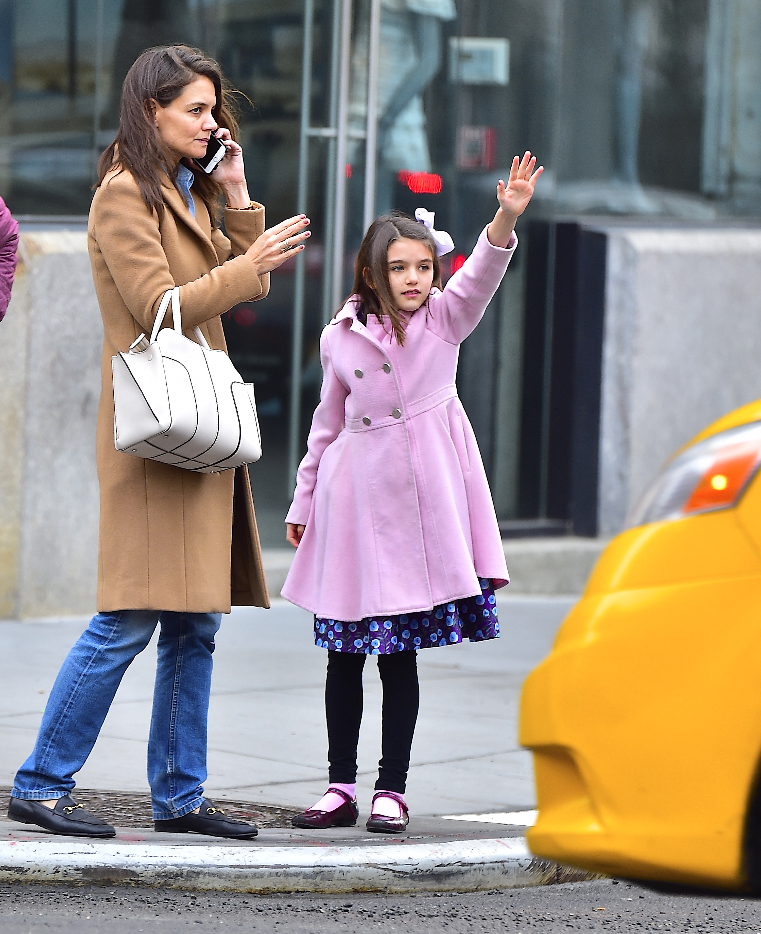 Suri Cruise and her mother, Katie Holmes, are captured standing curbside, patiently waiting for a taxi. Framed by the city's everyday hustle, the pair share a quiet moment amid the movement around them. Suri stands close by her mother’s side, their matching calm expressions offering a glimpse into their close-knit bond—even in the middle of a busy New York street.