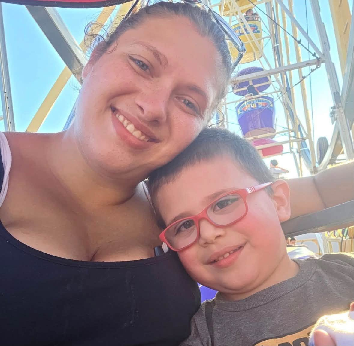 Metallica Rose Harrison cuddles up to her young son, who wears his distinctive red glasses, during a sunny outing beside a Ferris wheel. | Source: Facebook/TrueCrimeMama