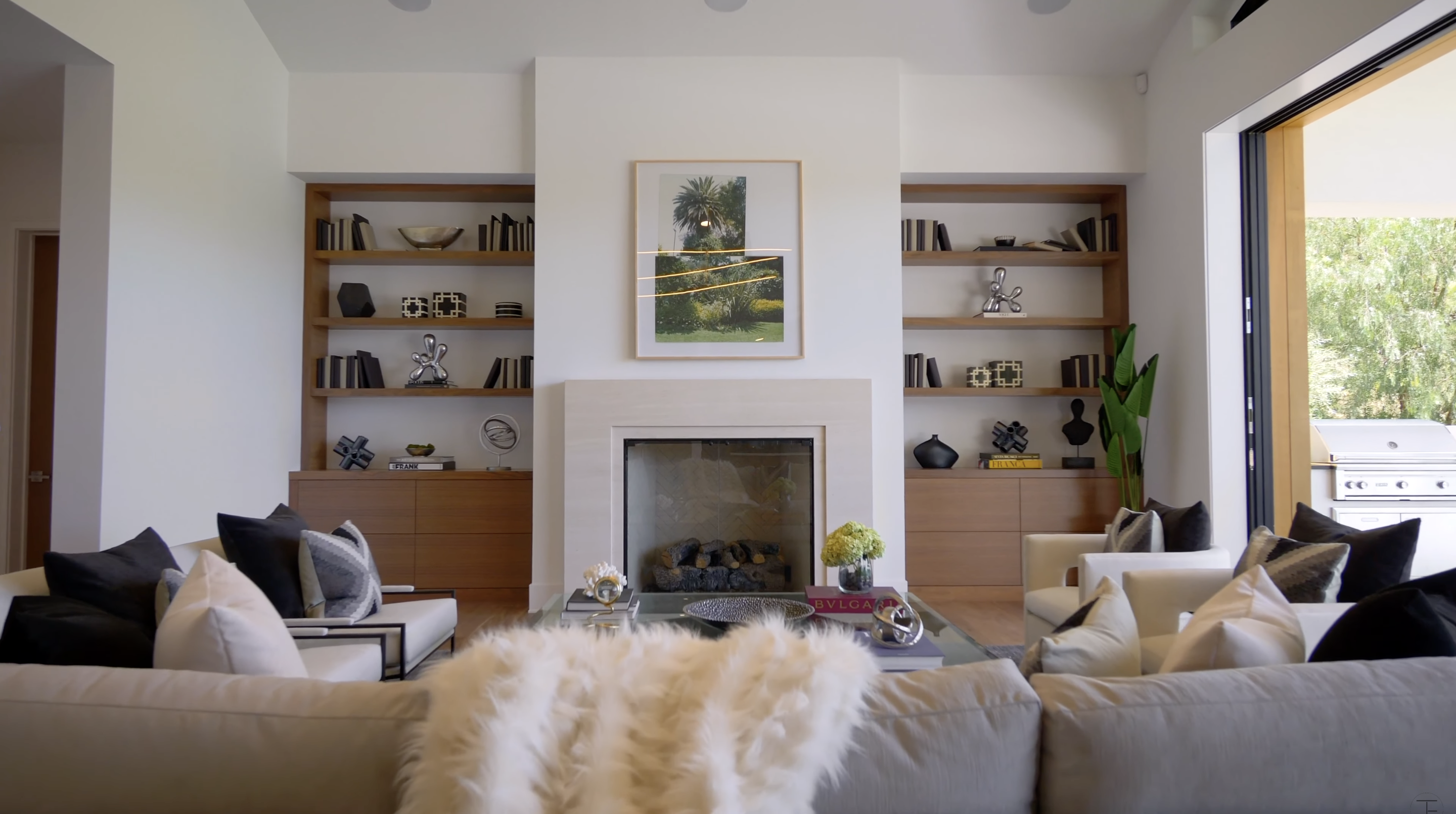 Wood cabinetry and shelving frame the fireplace in another sitting area. An opening beside the room leads toward an outdoor cooking space, placing the backyard only steps from the seating area. | Source: Youtube/Tomer.Fridman