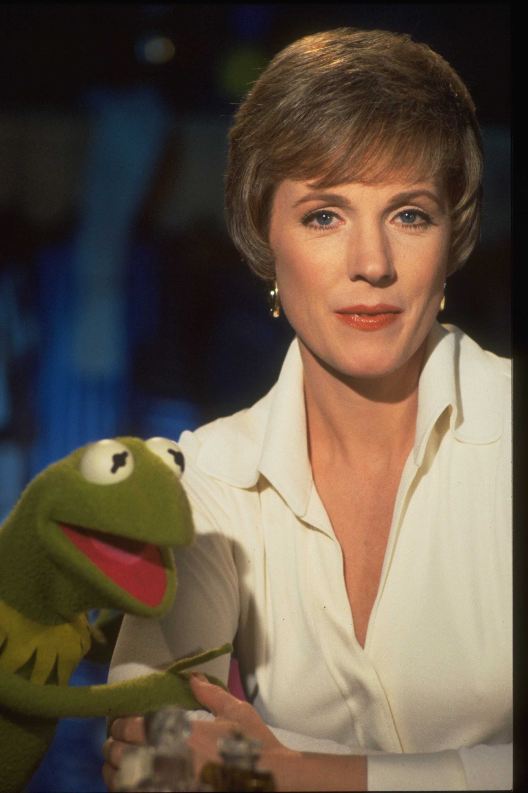 Julie Andrews shares a charming moment with puppet character Kermit the Frog on the set of "The Muppet Show" at Elstree Studios in Hertfordshire, circa 1977. The star looks chic and poised, gently holding Kermit's hand while wearing an open-collared white button-down shirt, subtle gold hoop earrings, and her signature side-swept feathered crop.