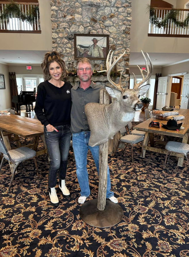 A towering stone feature anchors this large gathering room at the Governor's Residence. Long wooden tables, Western artwork, and a mounted deer fill out the space around it. | Source: Facebook/kristi.noem