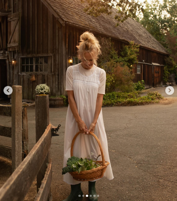 Pamela Anderson smiles softly as she carries a woven basket packed with freshly harvested garden greens in front of the rustic wooden barn at Arcady. Dressed in an airy white dress and green rubber muck boots, she channels the grounding simplicity of estate life while celebrating "Pamela's Cooking with Love," sharing gratitude for a show centered around her beloved Vancouver Island farm and garden. | Source: Instagram/pamelaanderson