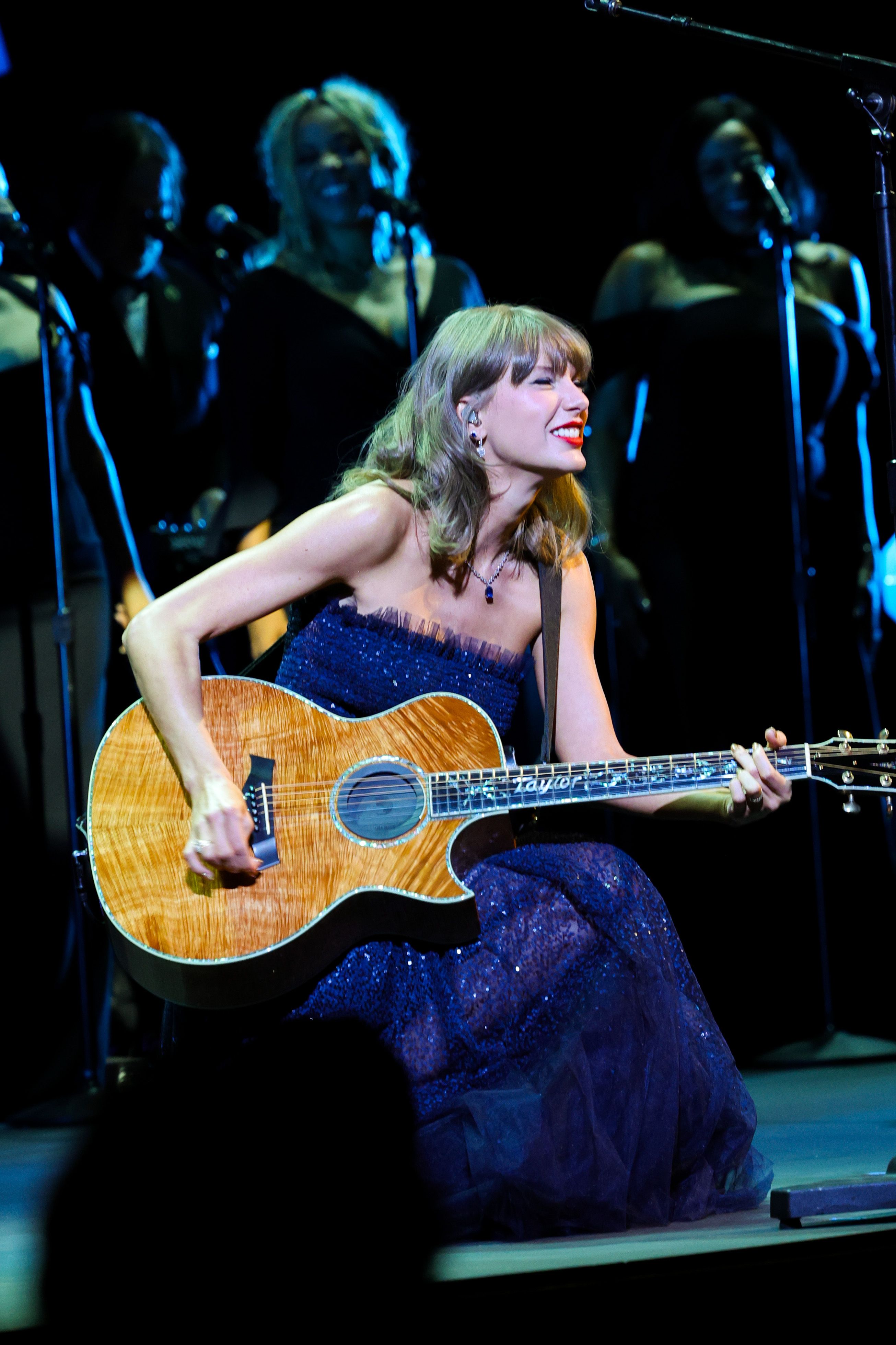 Taylor Swift performs an acoustic set in a strapless midnight blue gown shimmering with all-over sequins and delicate tulle trim along the neckline. Seated with her signature custom acoustic guitar, the singer pairs the sparkling stage look with a delicate sapphire pendant necklace, diamond drop earrings, her classic red lip, and soft honey-blonde waves.