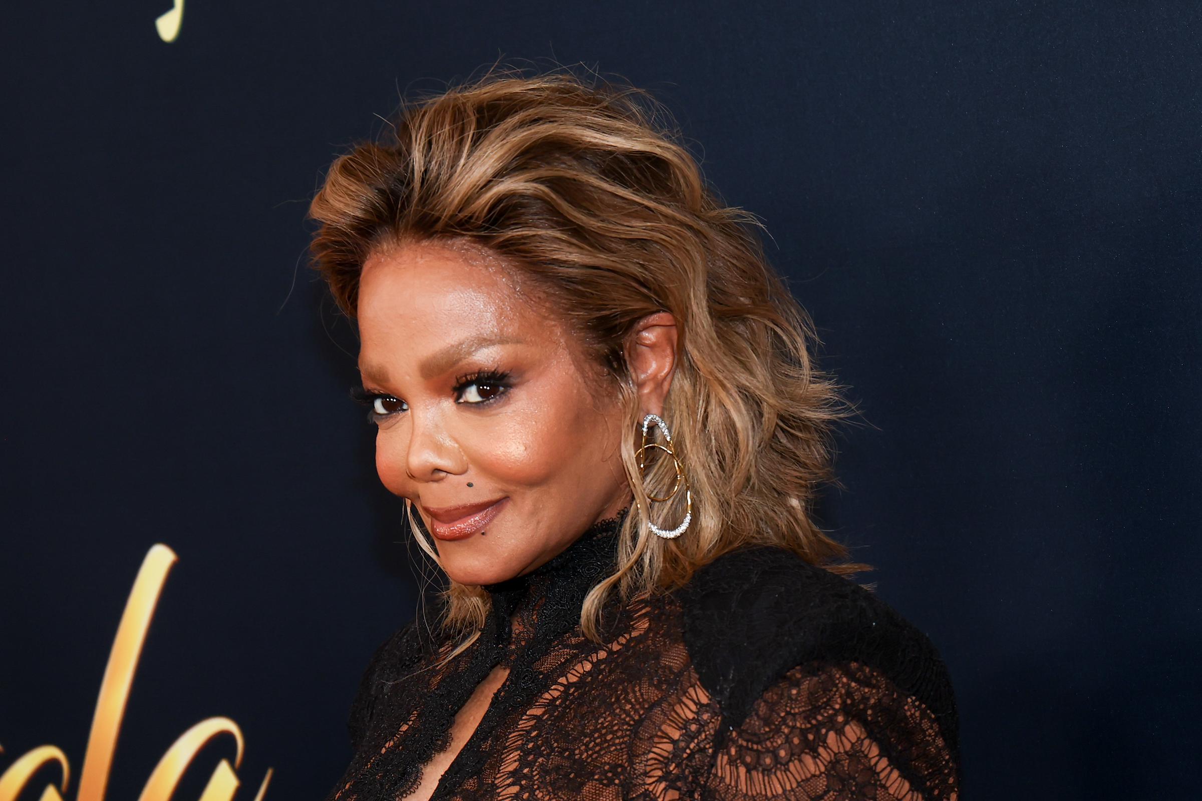 Janet Jackson poses for a close-up photo, highlighting her swept-back, highlighted hair, statement gold-and-silver hoop earrings, and the intricate floral lace and structured shoulders of her black gown. | Source: Getty Images