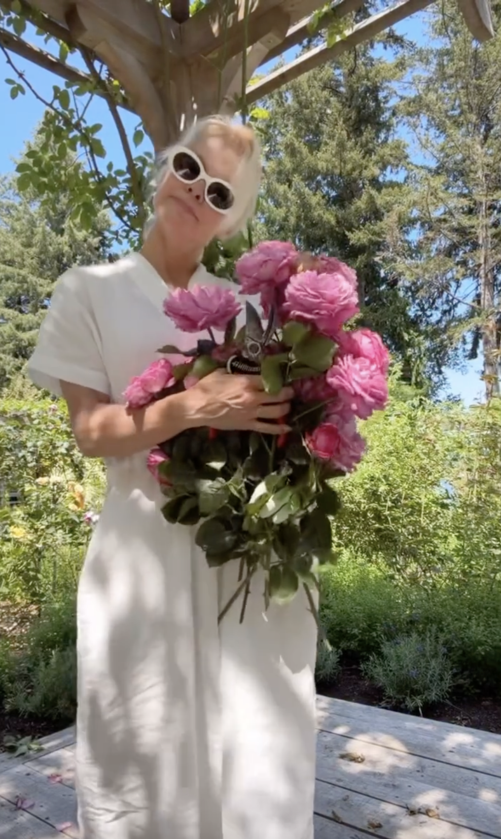 Pamela Anderson poses playfully beneath a garden arbor at Arcady, holding garden shears and an enormous bundle of freshly picked pink roses against her white linen dress. Wearing bold white oval sunglasses, she effortlessly embodies the joy of cultivating her own flowers, proudly showing off blooms harvested from her flourishing 15,000-square-foot sanctuary. | Source: Instagram/pamelaanderson