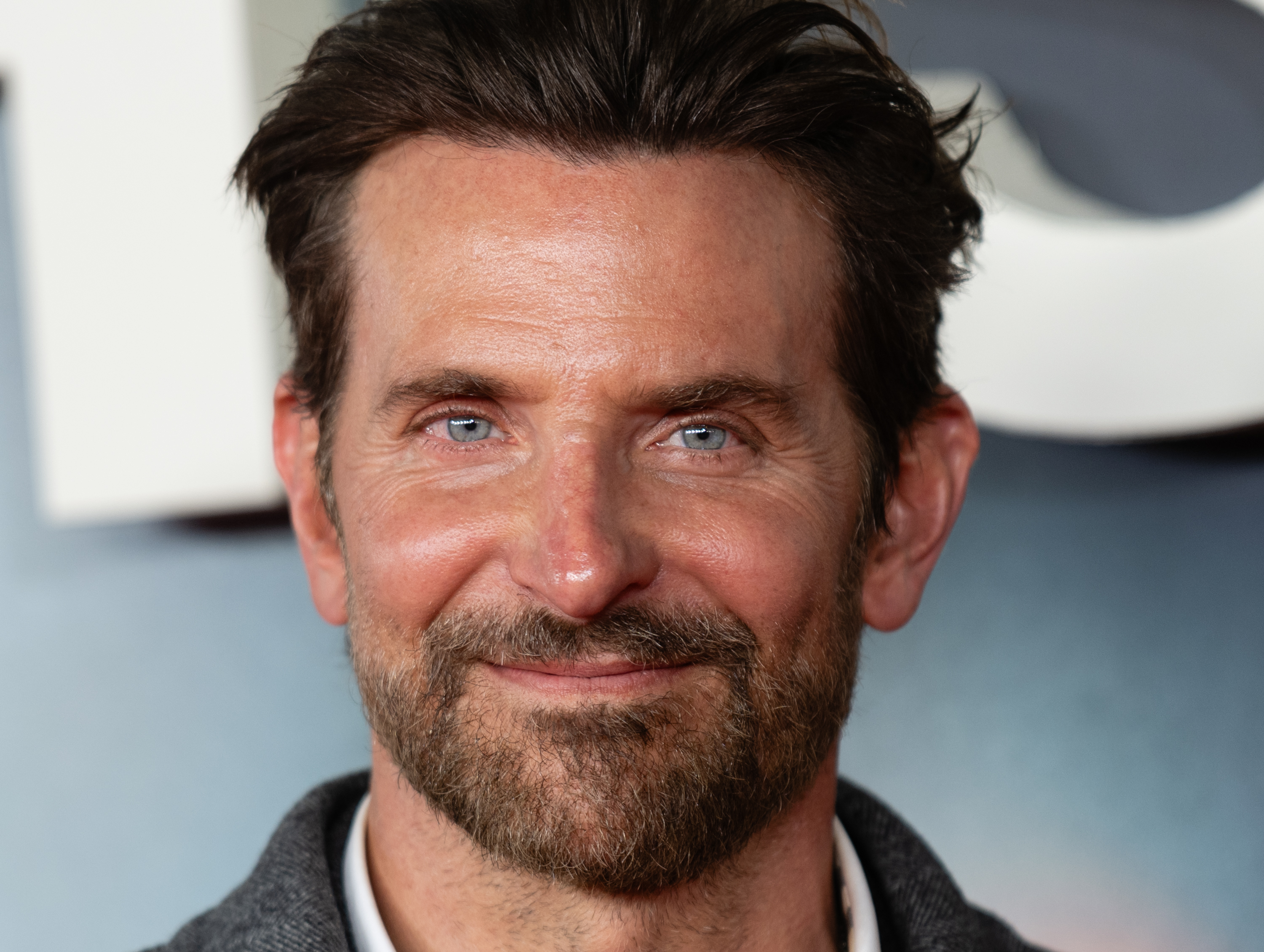 Bradley Cooper smiled warmly for a close-up portrait while attending the UK gala screening of "Is This Thing On?" at Odeon Luxe West End on January 19, 2026, in London, England. The actor showed off a neatly groomed beard and mustache alongside his swept-back hairstyle, dressed in a textured grey jacket layered over a crisp white collared shirt. | Source: Getty Images